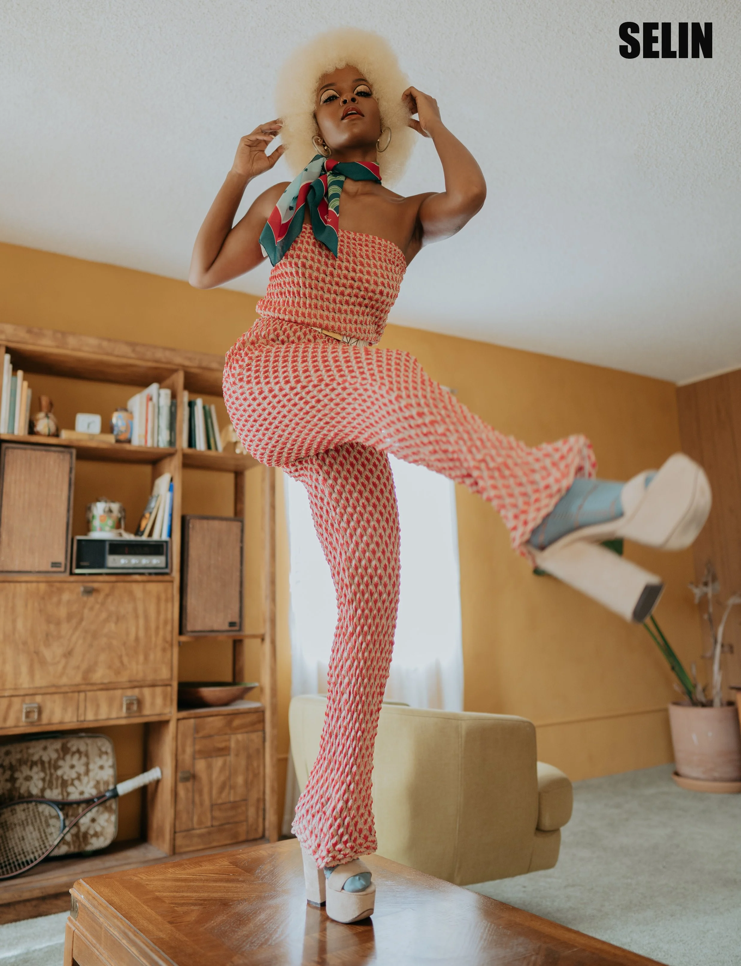 A woman with blonde afro hair dancing on a wooden table in a living room with yellow walls, wooden shelves, and a beige chair.
