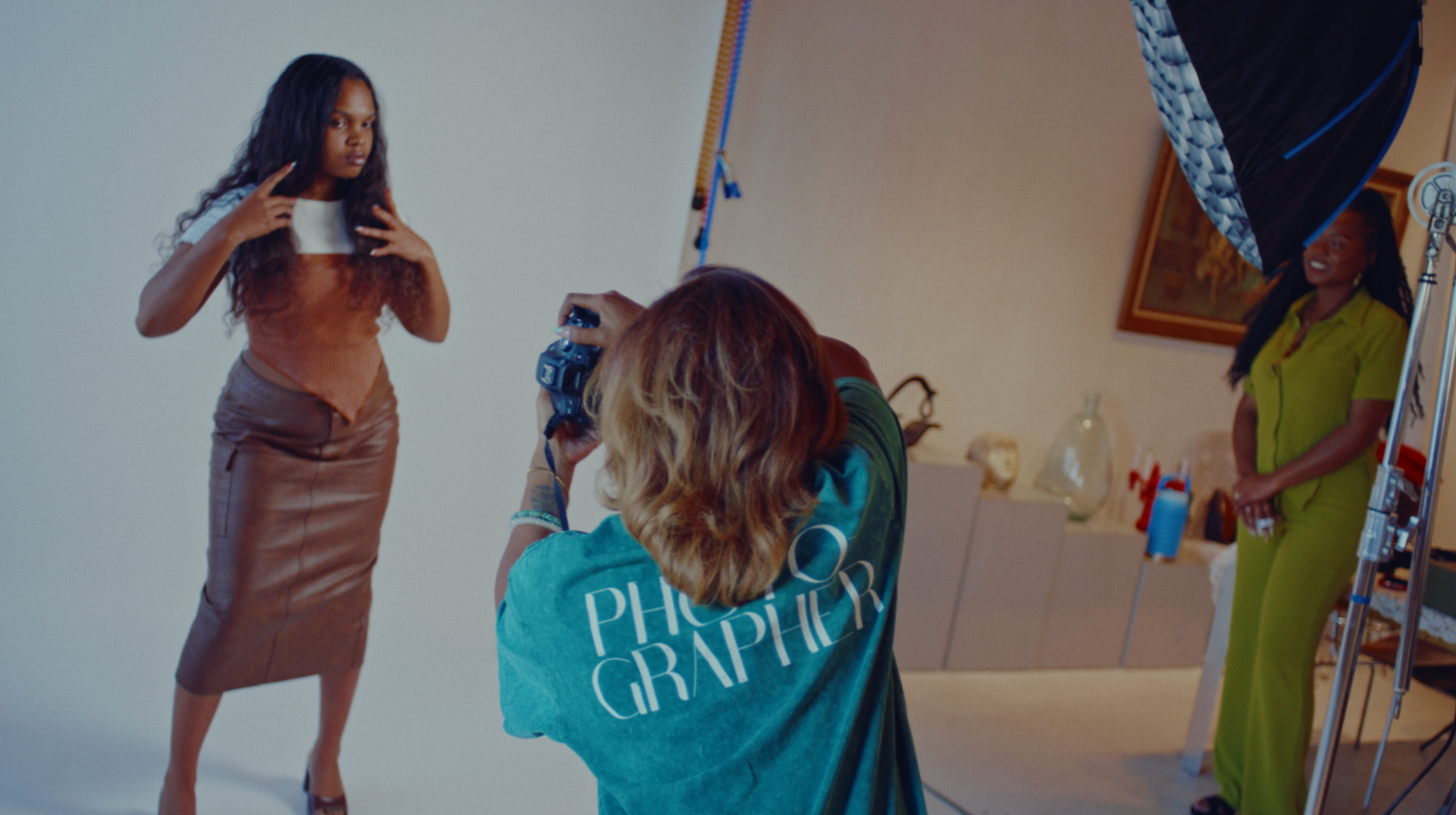 A woman in a shiny brown outfit poses during a photo shoot with a photographer taking her picture and a model in a green outfit smiling in the background.