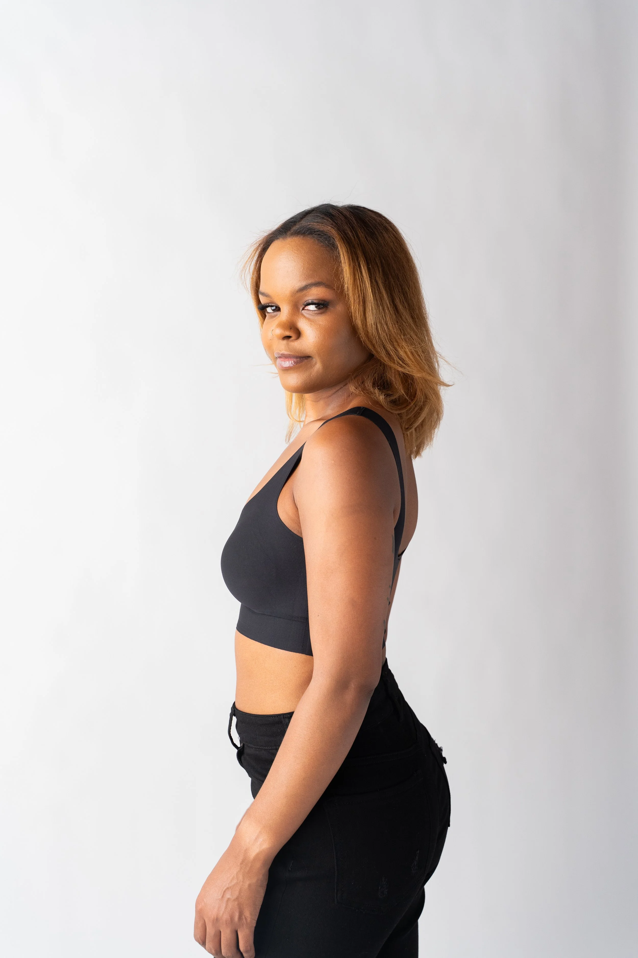 A woman with shoulder-length light brown hair, wearing a black crop top and black pants, is standing against a plain white background looking at the camera.