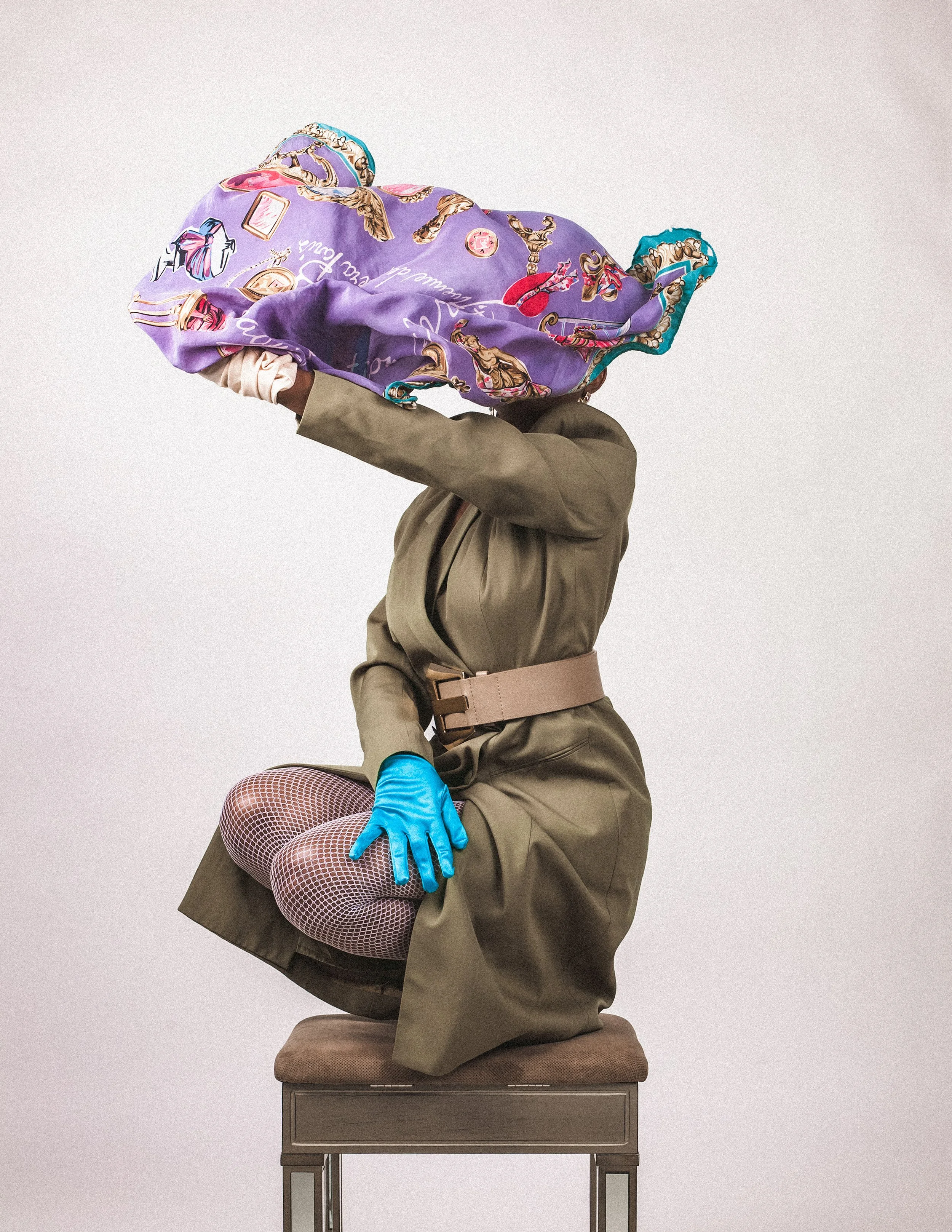 A person kneeling on a chair, wearing fishnet stockings, a khaki dress with a wide belt, and blue gloves, with their head covered by a colorful silk scarf.