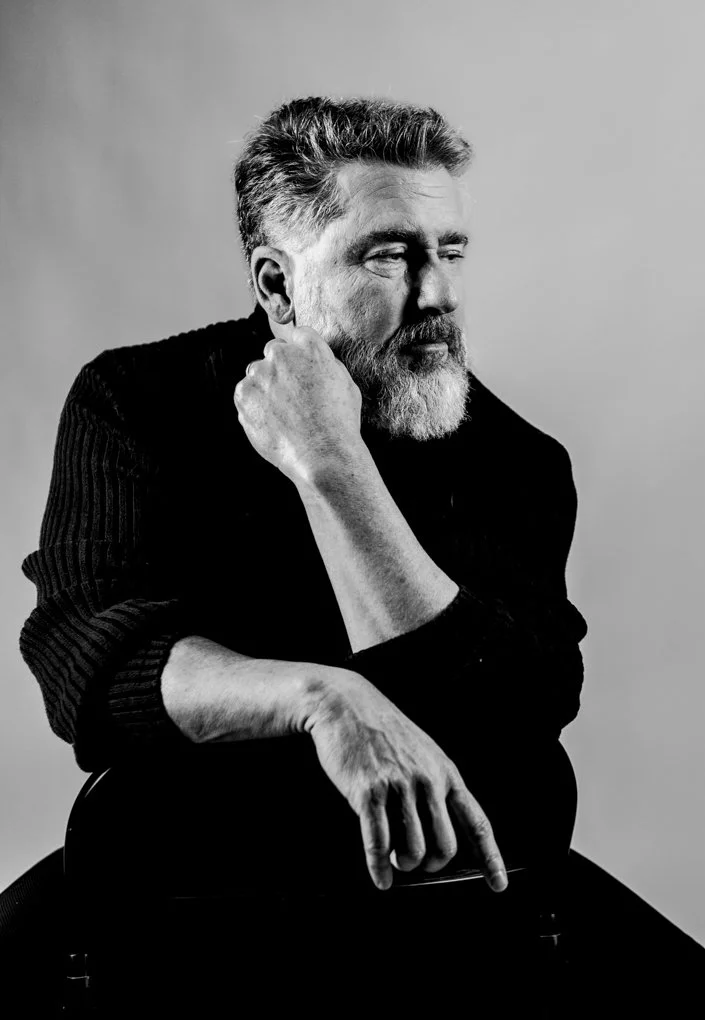 Black and white portrait of a middle-aged man with a beard and mustache, wearing a dark sweater, sitting with his hand resting on his face and looking thoughtfully to the side.