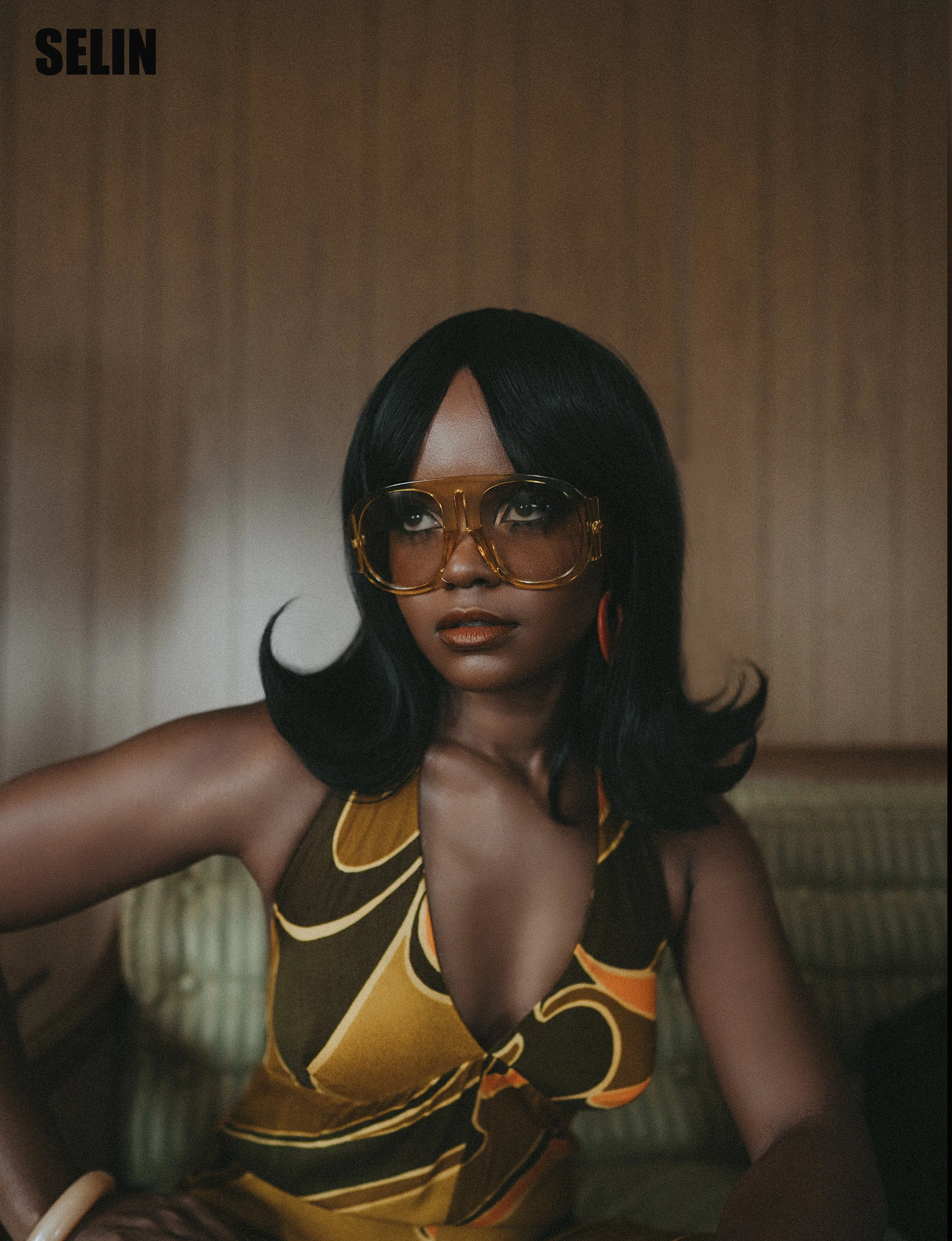 A woman with black hair wearing large yellow sunglasses, orange earrings, and a colorful dress with brown, yellow, and orange patterns, sitting on a green couch with a wooden wall background.