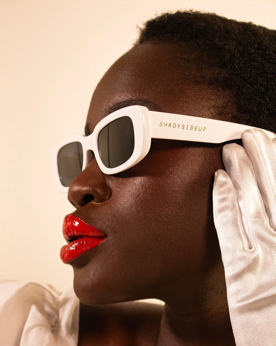 Close-up profile of a woman wearing white sunglasses with gold lettering, red lipstick, white glove, and a white top against a plain background.