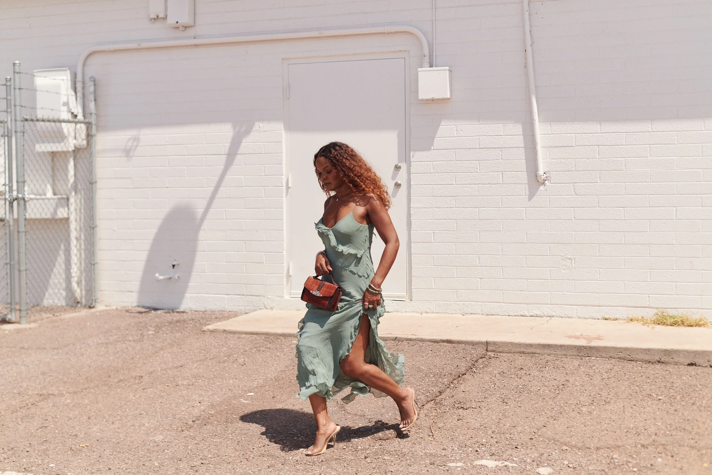 A woman in a flowing green dress with ruffles and a slit, holding a small handbag with a tiger pattern, walking barefoot on a sunny day near a white brick wall and a chain-link fence.