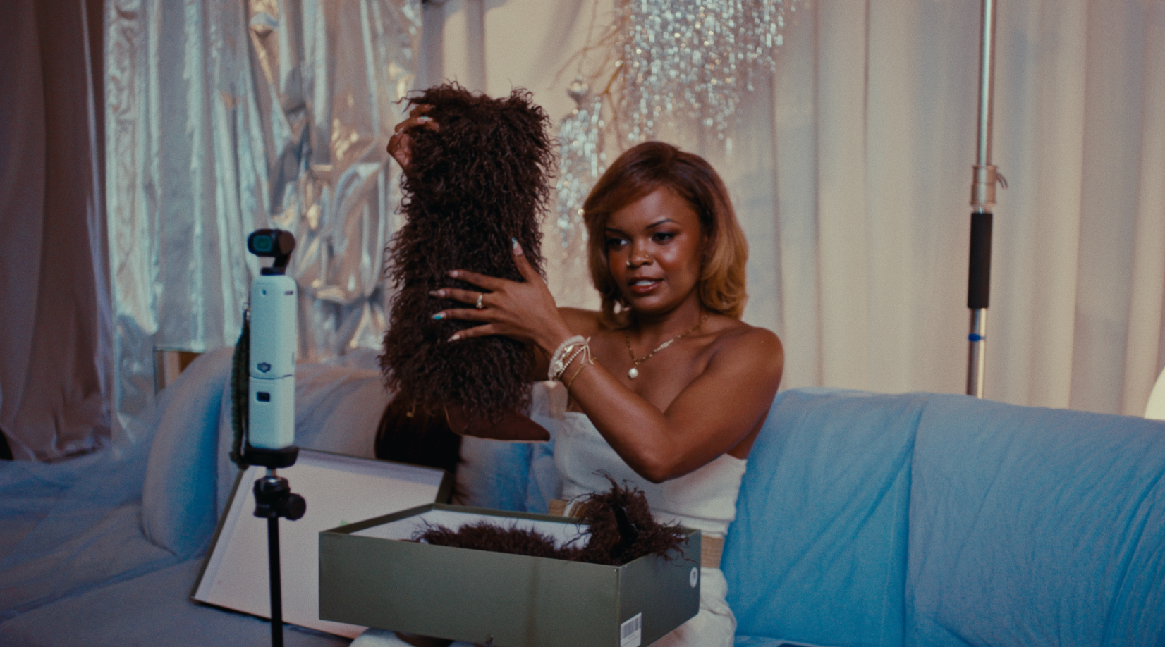 A woman sits on a couch holding a mannequin head with curly hair. There is a box of hairpieces or wigs on her lap and a mirror in front of her. The background features a decorative, shiny curtain and dim lighting, indicating an indoor setting.