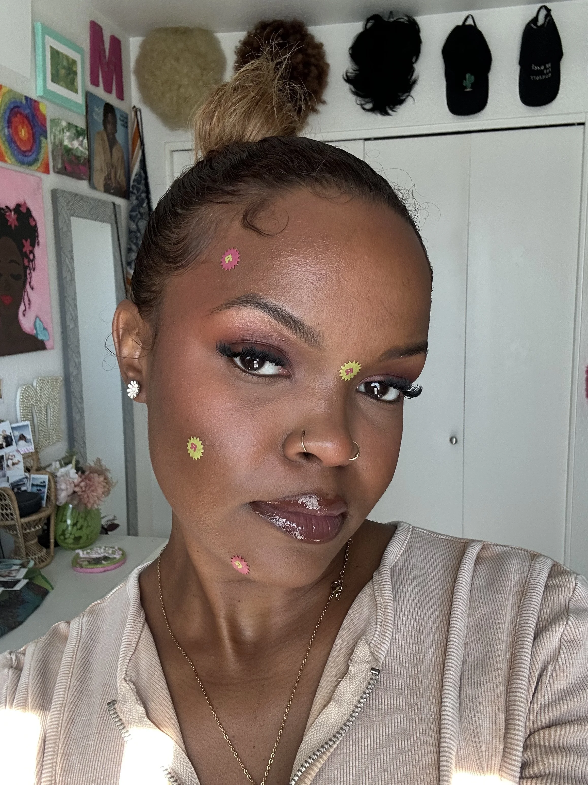 Close-up of a woman with styled hair, makeup, and decorative stickers on her face, standing in a room with various wall decorations and a wardrobe in the background.