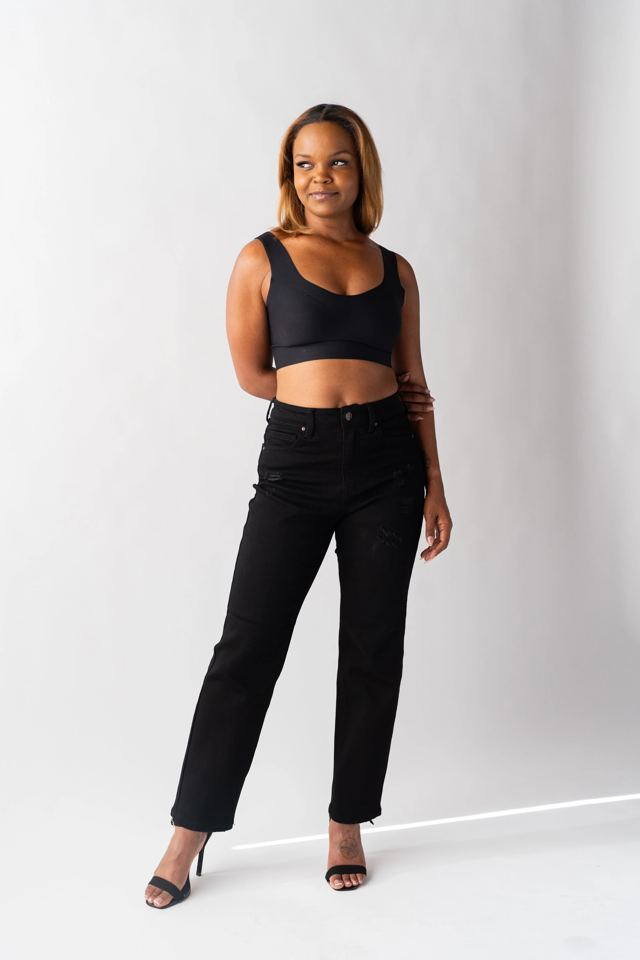 A woman with medium-length hair, wearing a black crop top, black ripped jeans, and high heels, standing against a plain white background.