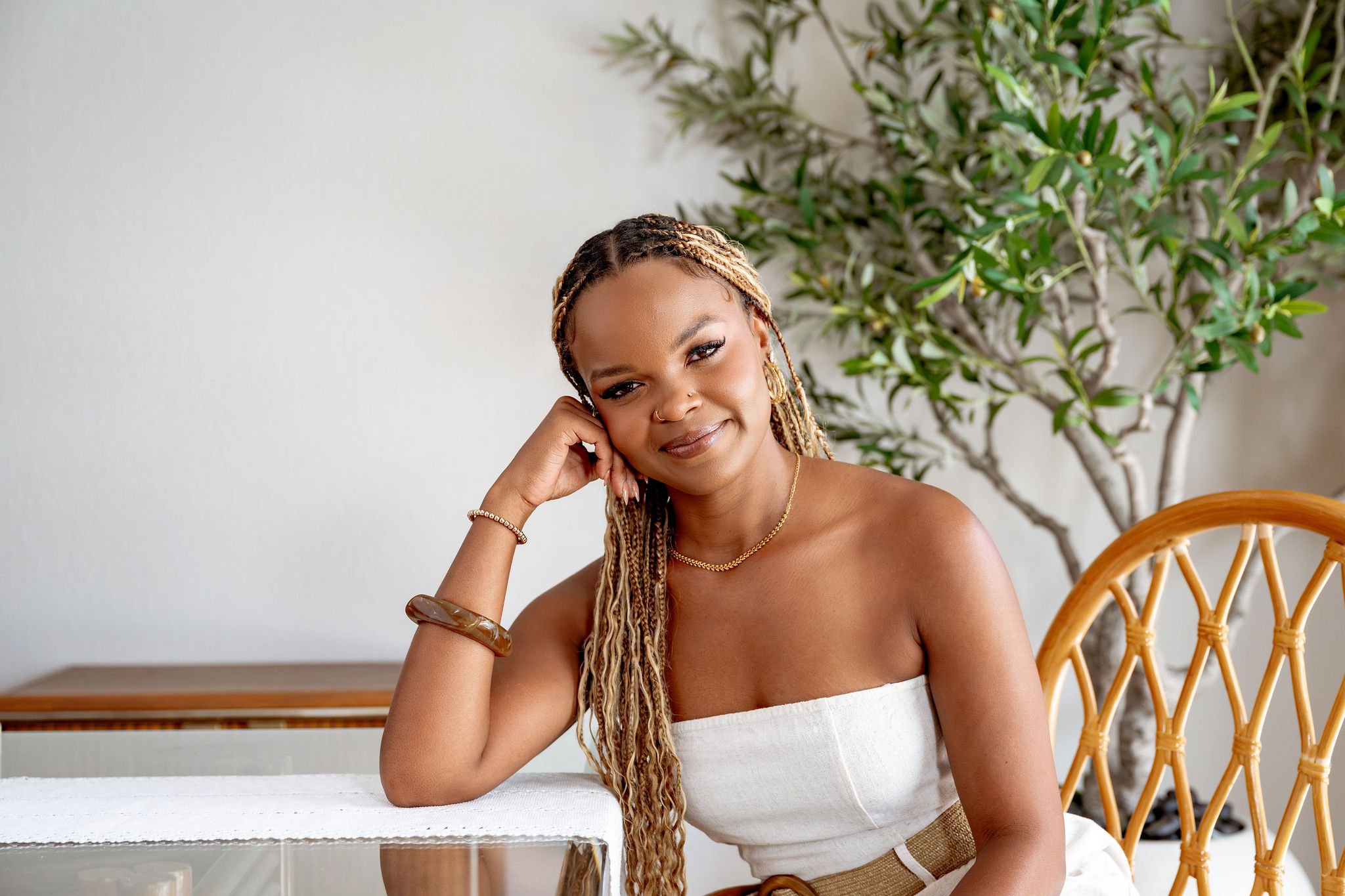 A woman with braided hair sitting at a table with a relaxed pose, resting her head on her hand, smiling softly, with a large plant and a wooden chair in the background.