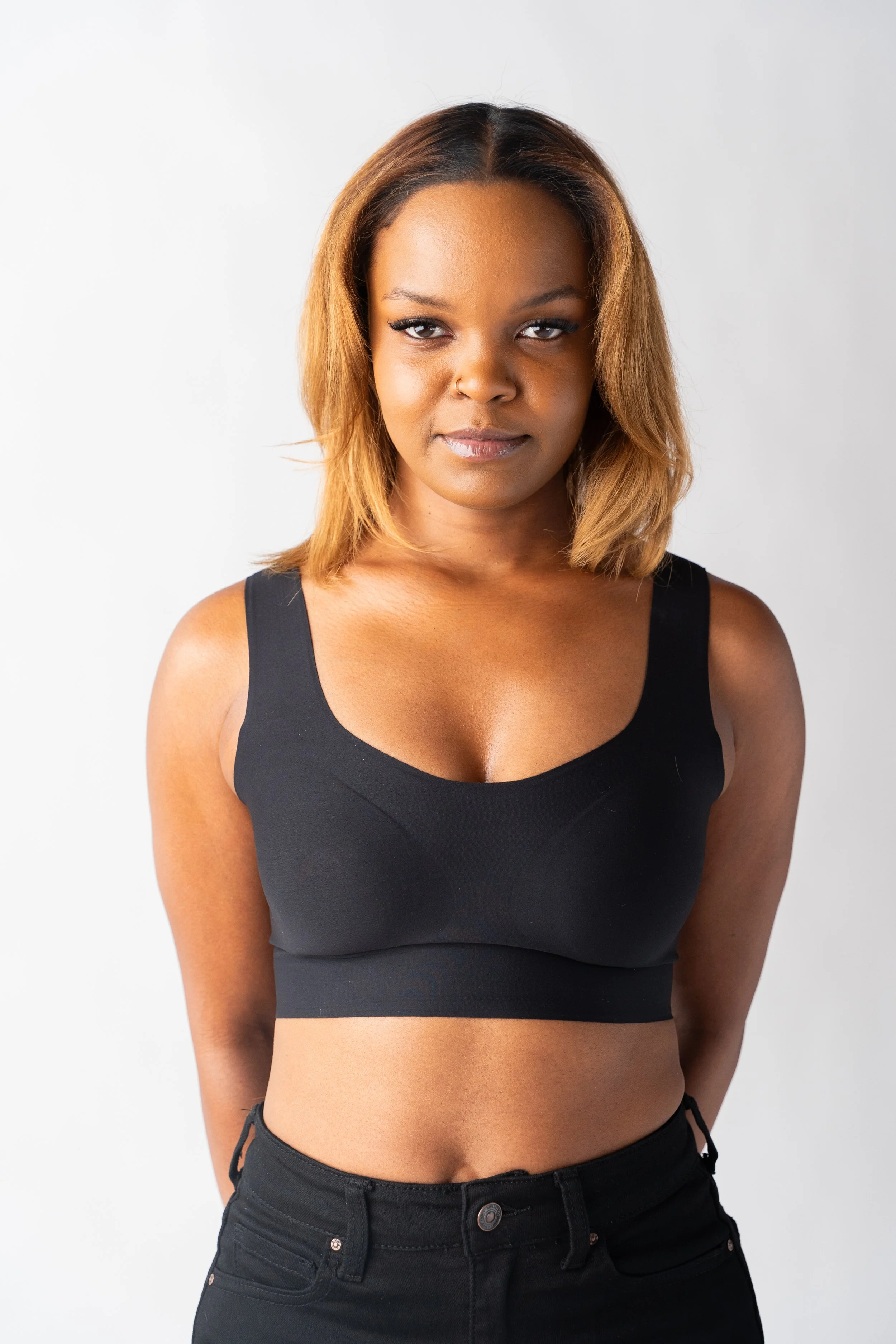 Portrait of a confident young Black woman with shoulder-length hair, wearing a black sports bra and black pants, standing against a plain white background.