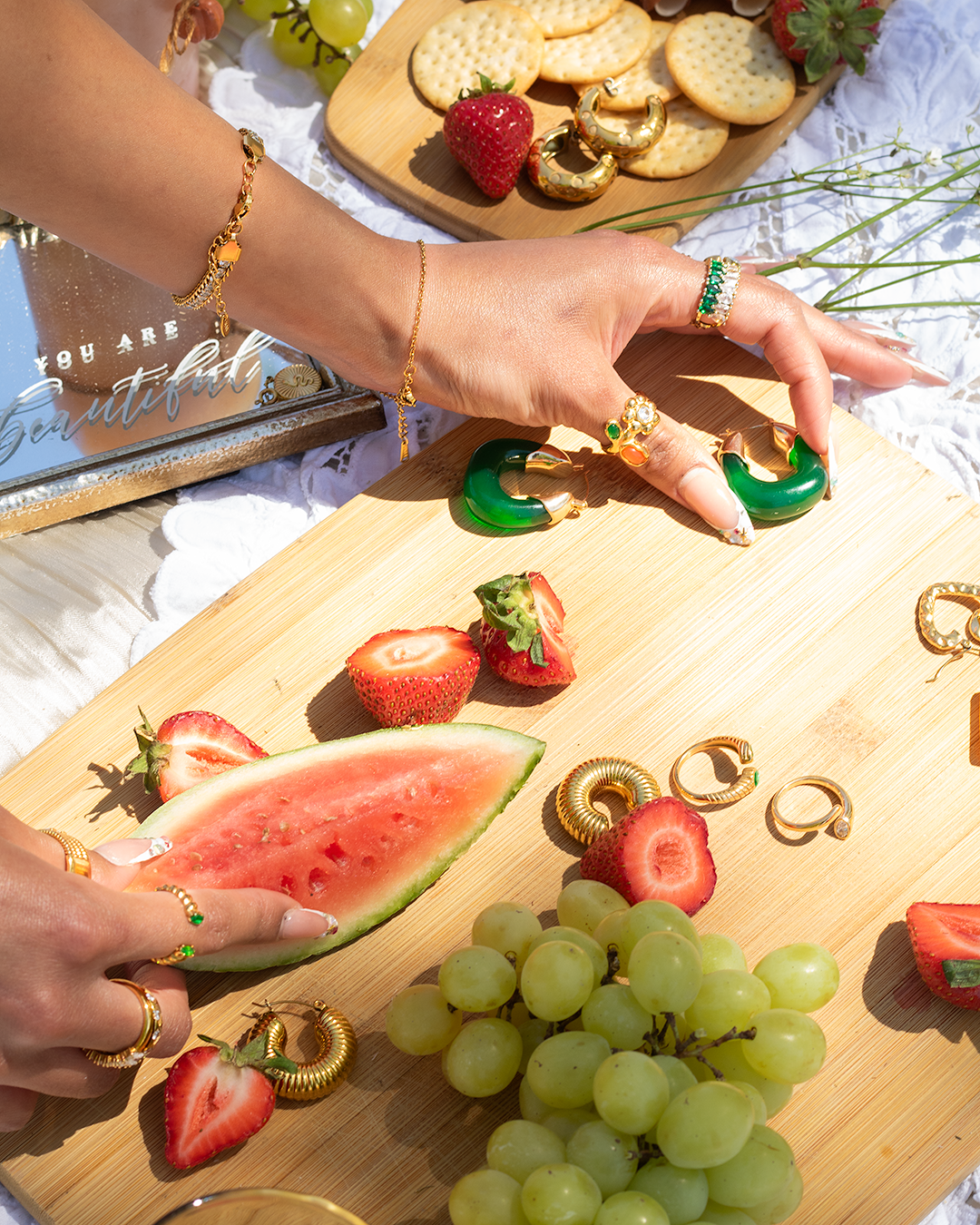 A person's hand with jewelry reaching for green watermelon-shaped earrings on a wooden cutting board. The board has strawberries, grapes, and watermelon, with gold rings and jewelry scattered around. A small wooden board with crackers, a strawberry, 