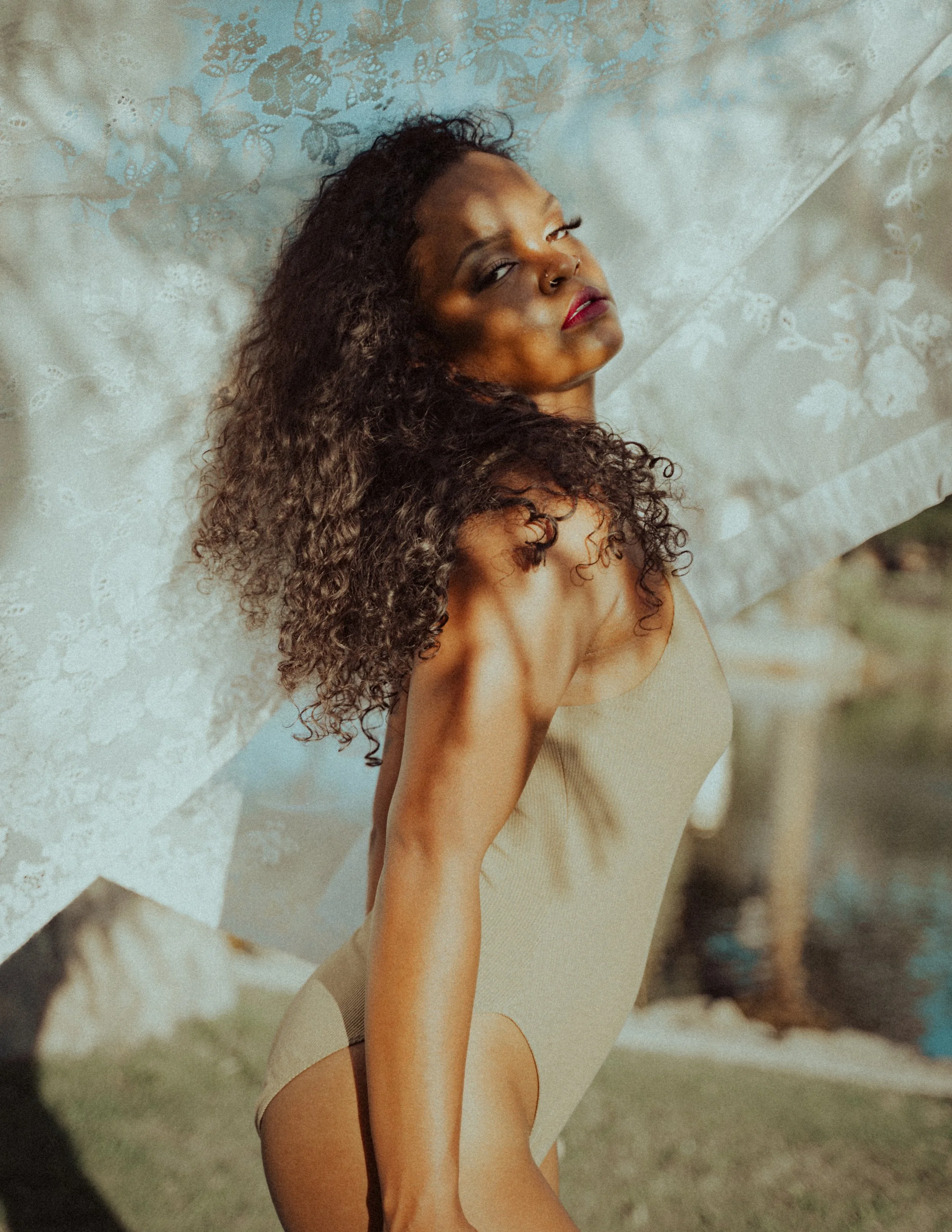 A woman with curly hair posing outdoors, leaning supported by a wall with lace curtains, wearing a beige bodysuit.