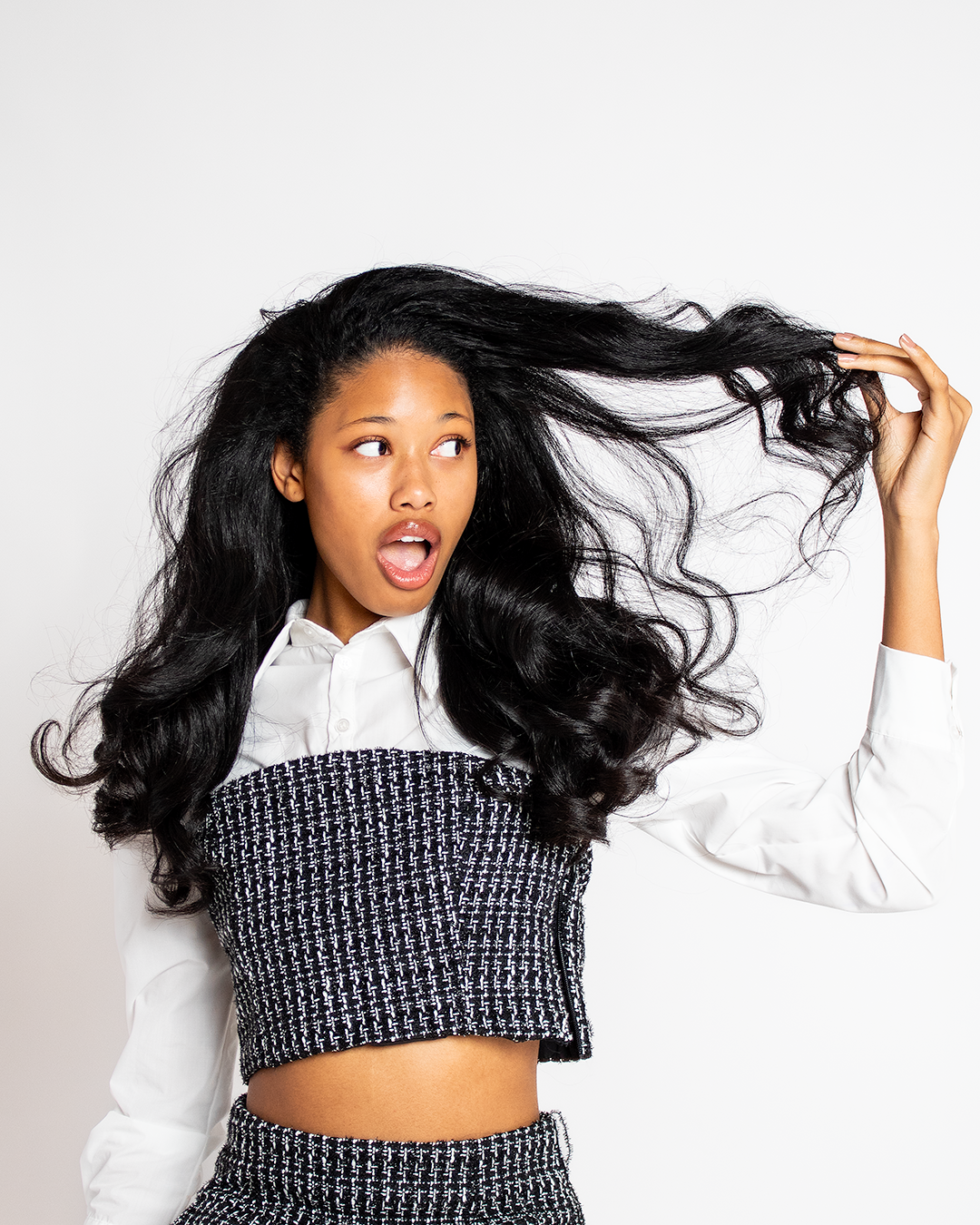 Young woman with long, curly black hair wearing a white shirt with a black and white patterned cropped vest, holding her hair while looking surprised or curious against a plain white background.