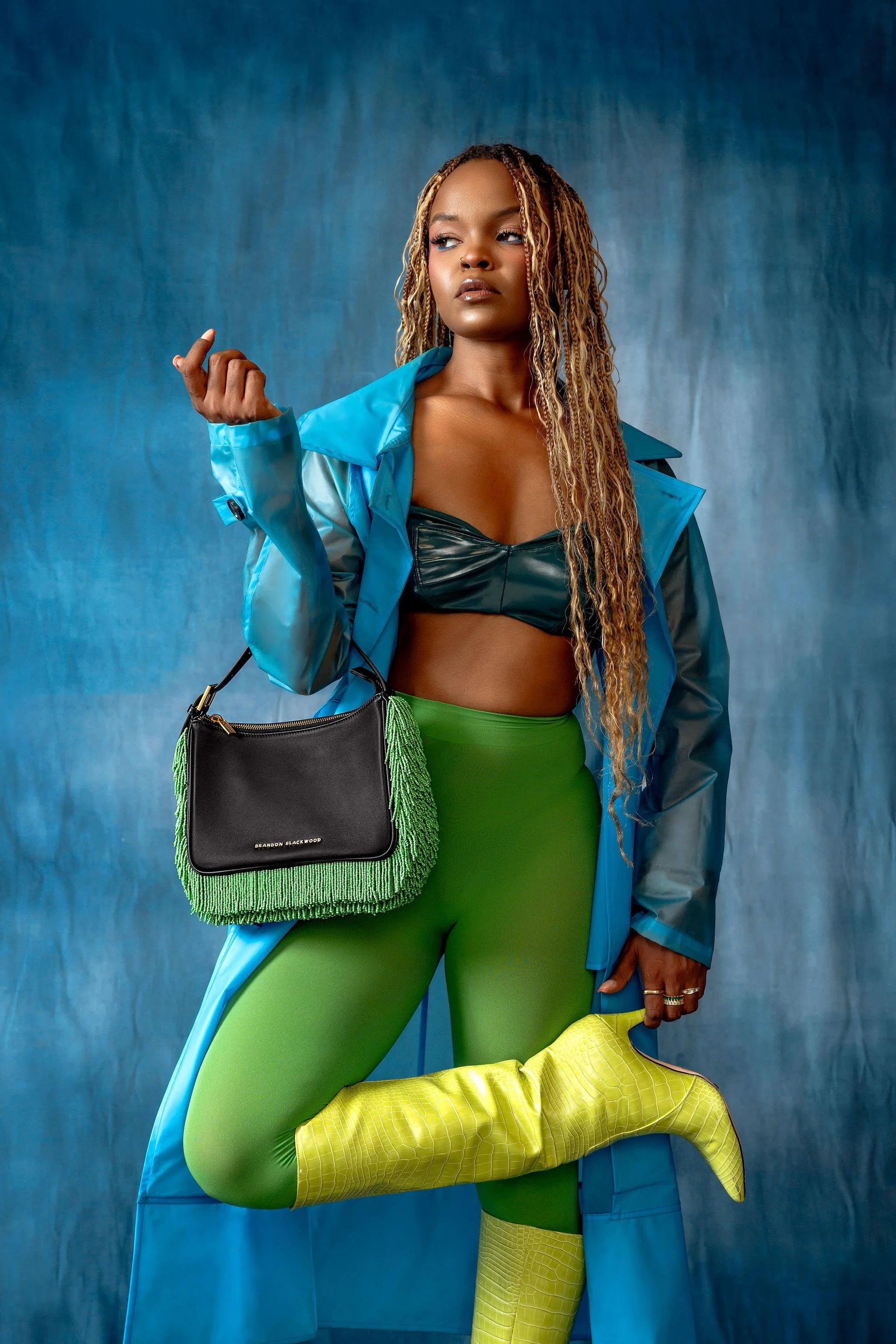 A woman with long curly hair wearing a blue raincoat, a black leather crop top, bright green tights, and yellow boots, holding a black and green fringe handbag, standing against a blue backdrop.