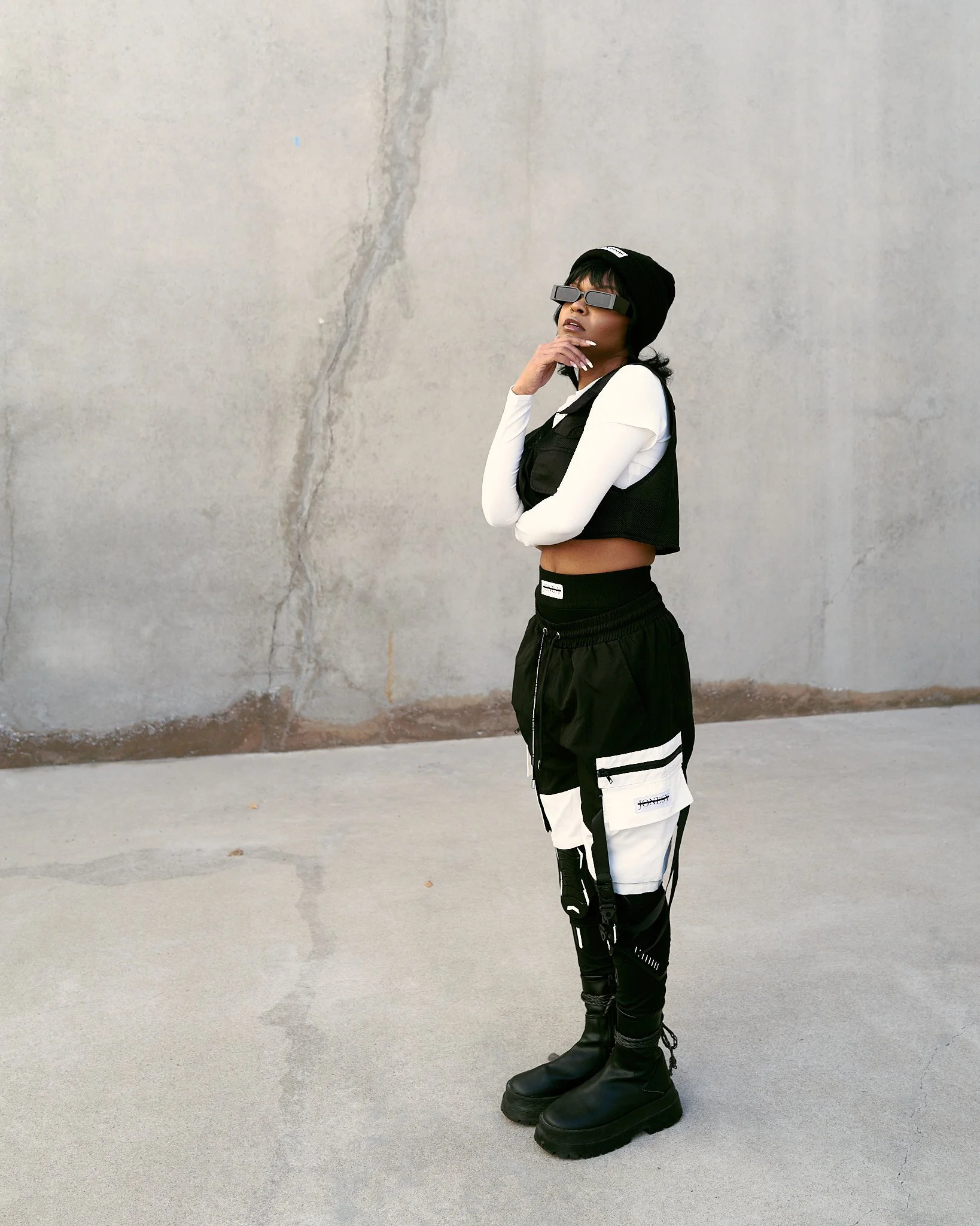 A woman stands against a concrete wall wearing black and white streetwear, including cargo pants, a cropped vest, and large boots, while posing with her hand on her chin.