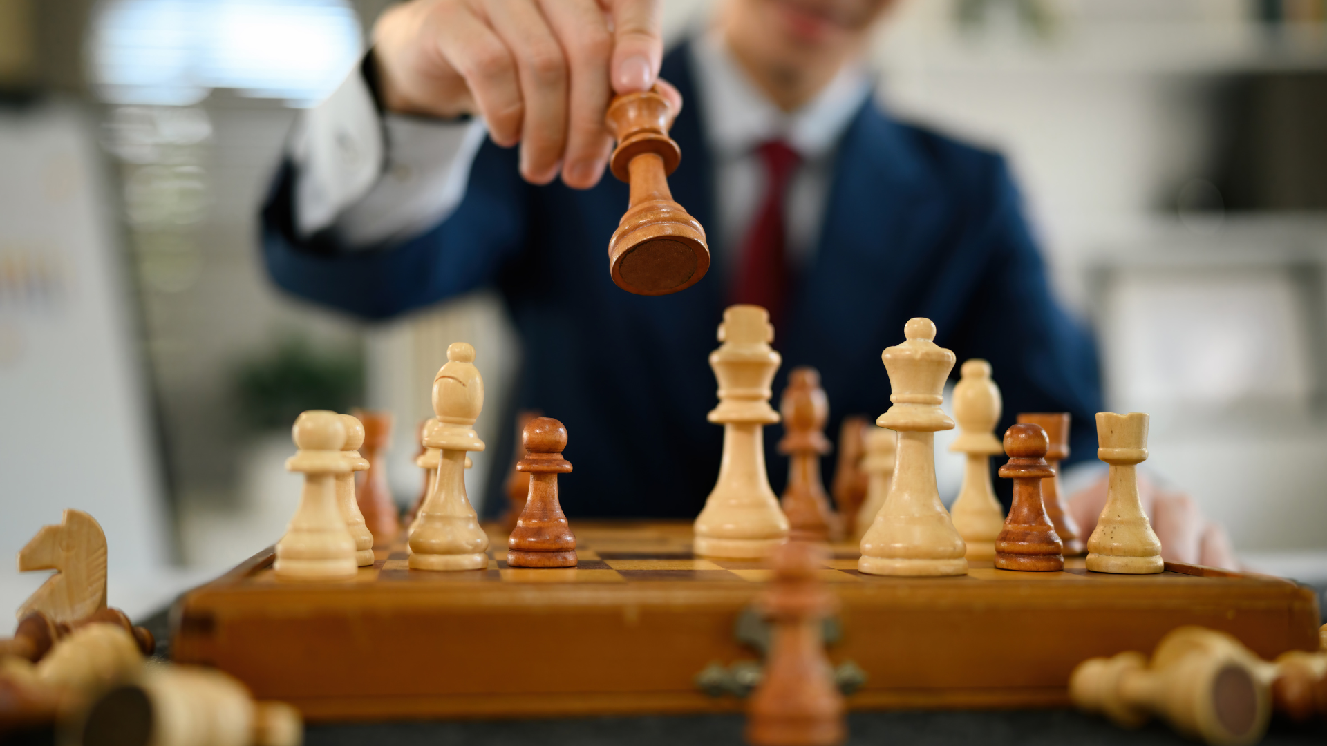 A person in a dark suit makes a move in a chess game, with chess pieces on a wooden board in focus.
