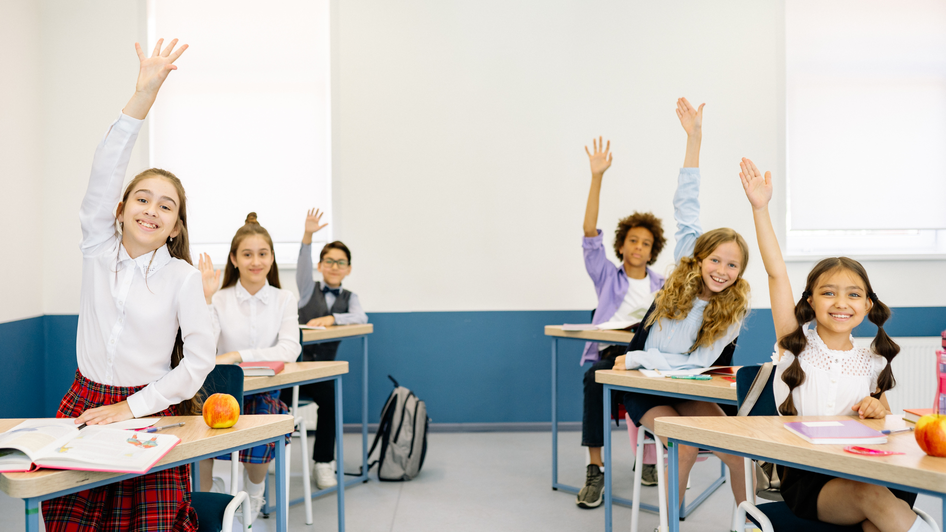 Classroom with six children sitting at desks, raising hands, with apples and books on desks, in a bright room with white and blue walls.