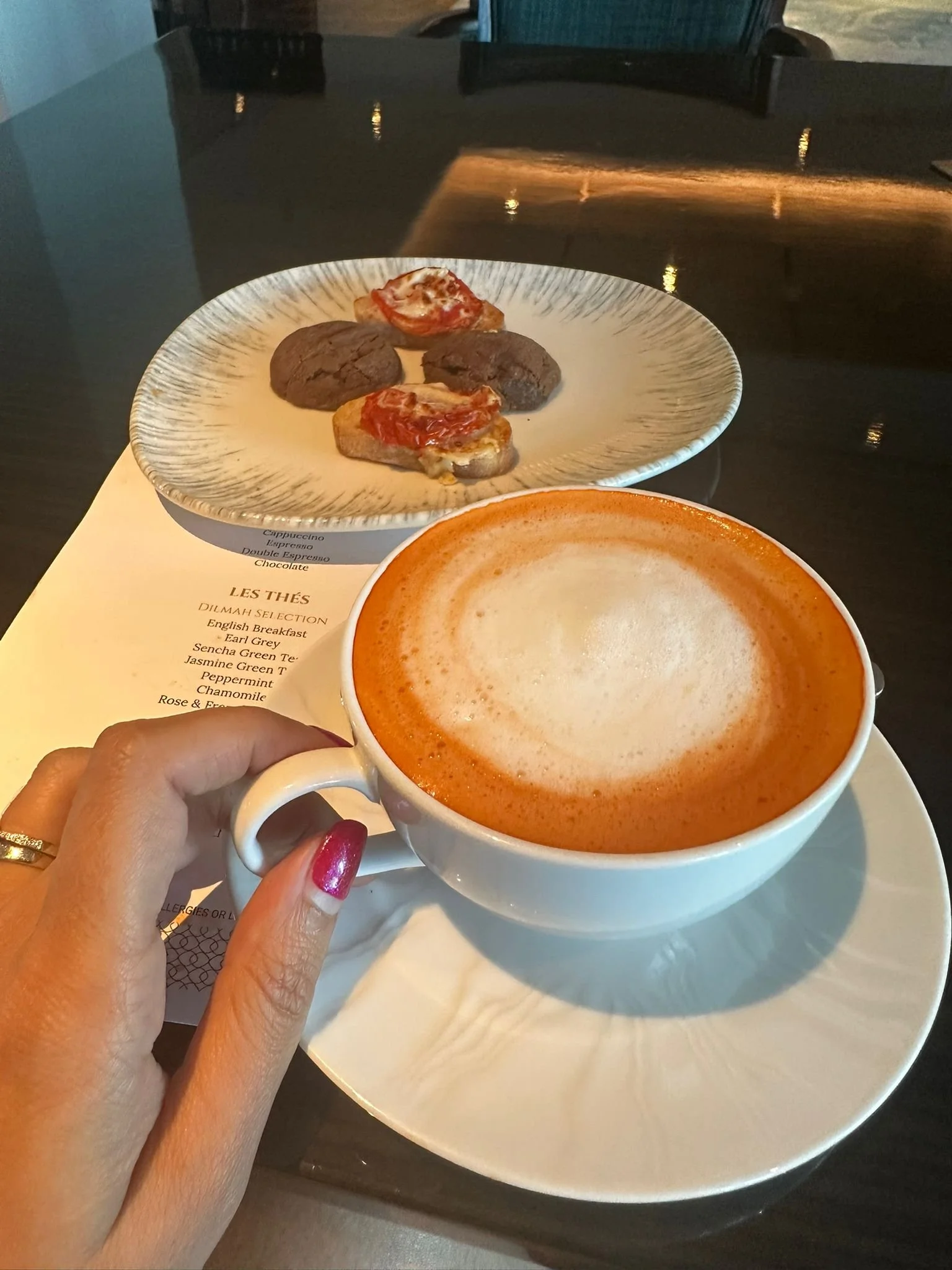 A hand holding a cup of latte with foam art, in the background a plate with four small cookies topped with cherry and tomato, and a menu partially visible.