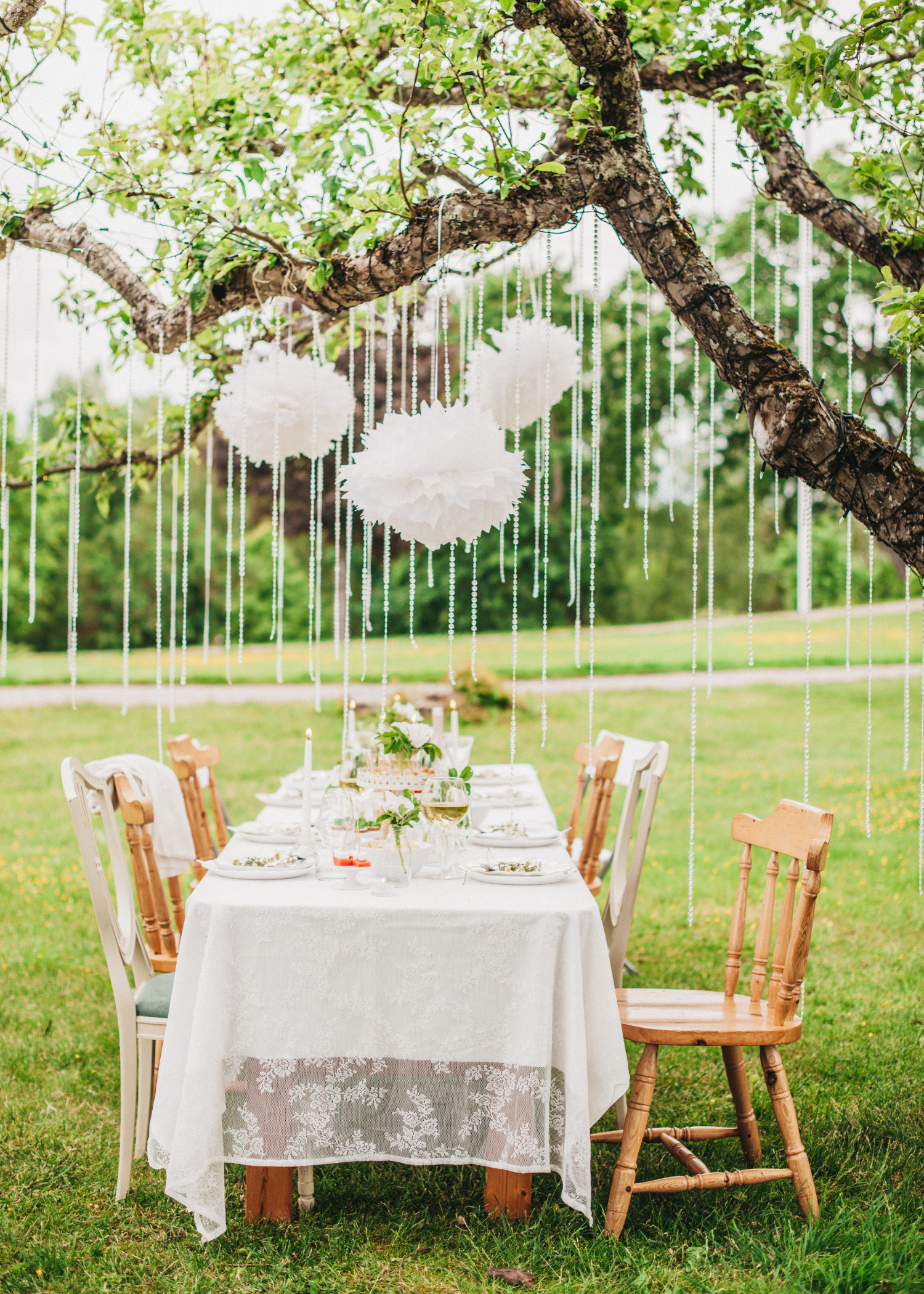 Outdoor dining table decorated with white lace tablecloth, candles, and green floral arrangements under hanging white paper pom-poms and decorations, set on grass beside a tree with a park or garden in the background.