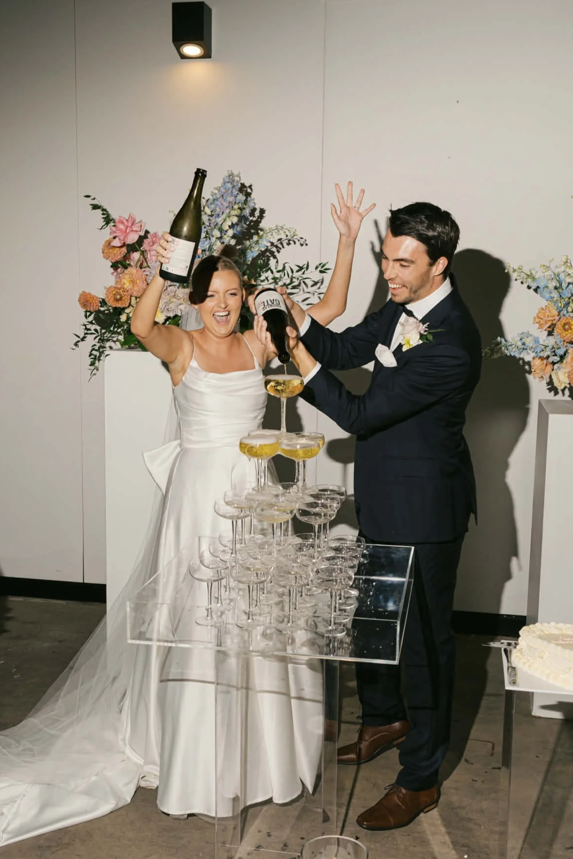 A bride and groom celebrating at their wedding with champagne, standing behind a tower of champagne glasses, with floral arrangements in the background.