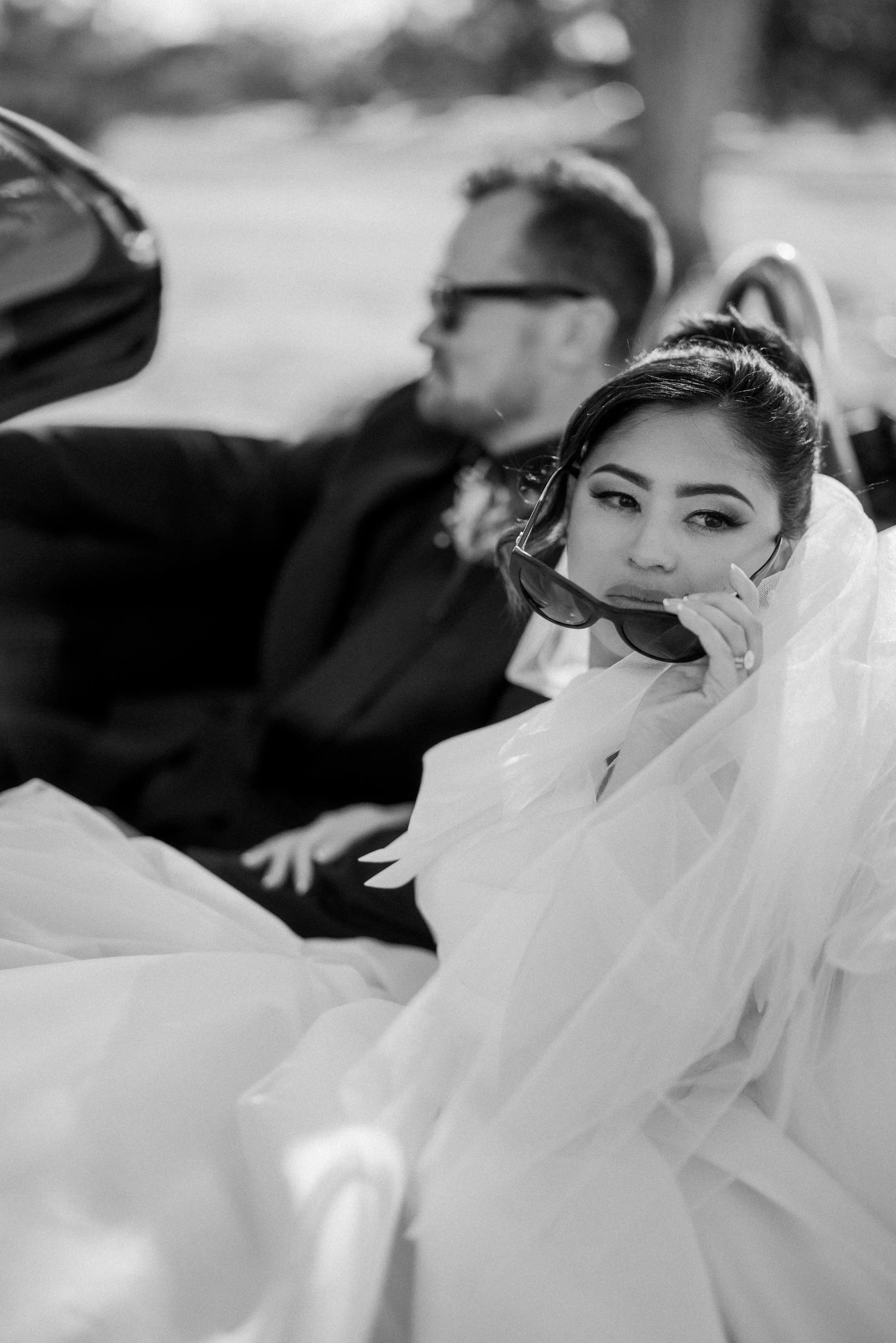 A woman in a wedding dress holding sunglasses, sitting in a car with a man in a suit in the background.