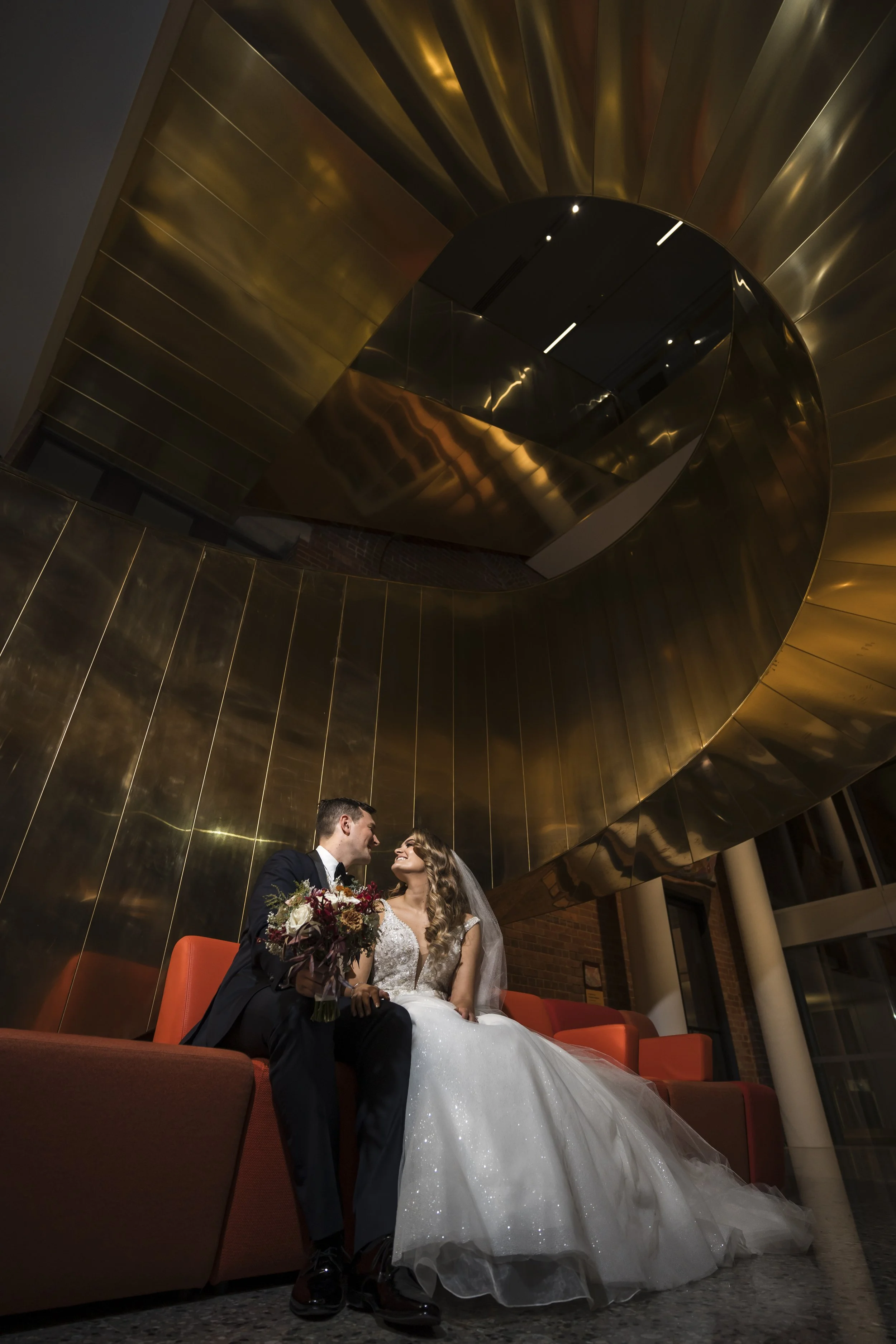 A bride and groom sitting on a couch in a modern, stylish lobby with a large gold spiral staircase overhead, looking into each other's eyes.