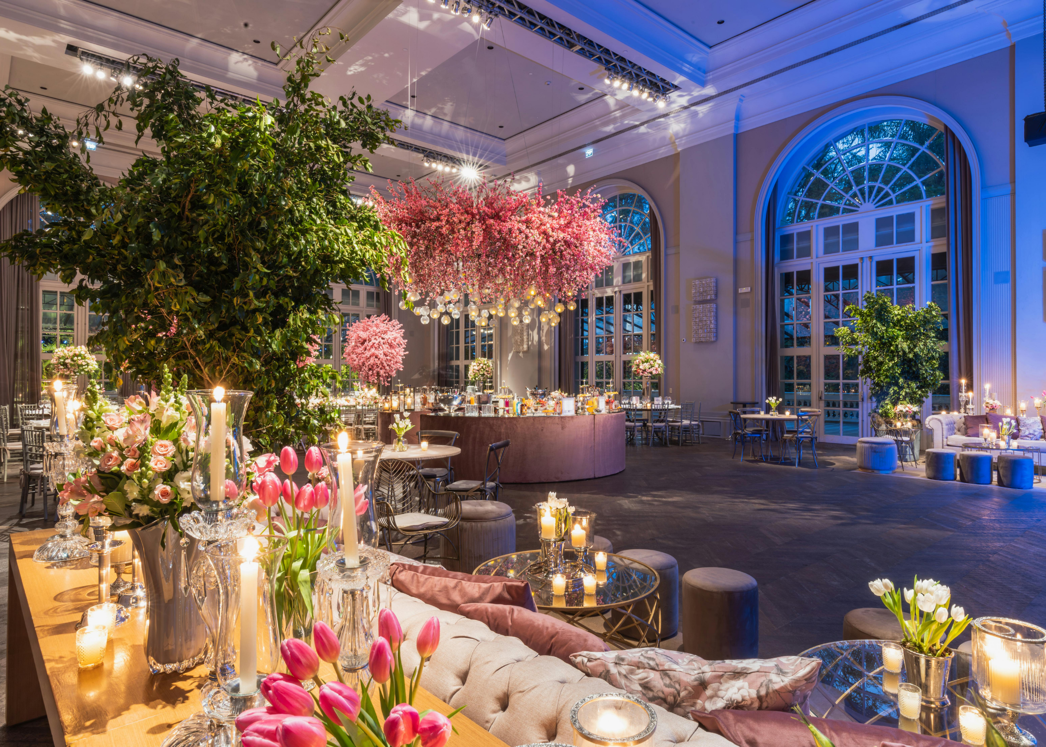 Elegant event space decorated with pink and white flowers, large hanging pink blossoms, and candles for an event.