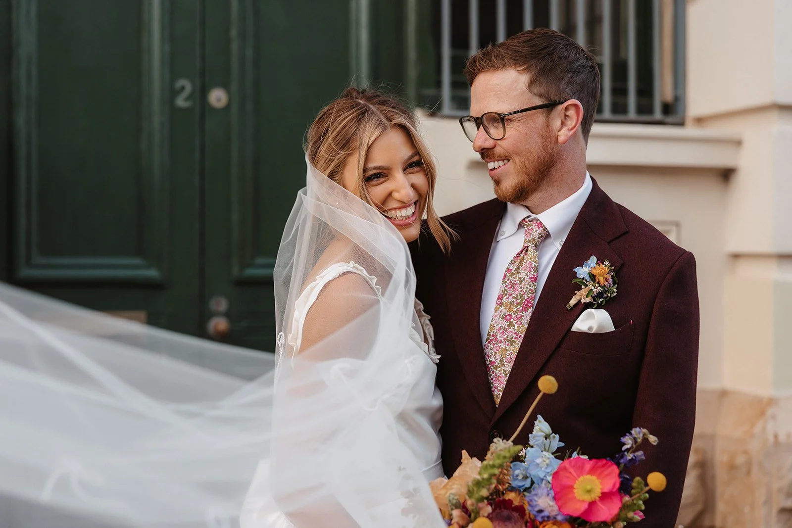 A smiling bride and groom on their wedding day, outdoors, with the bride in a white wedding dress and veil, holding a colorful bouquet, and the groom in a dark suit with a floral tie and boutonniere, sharing a loving moment.