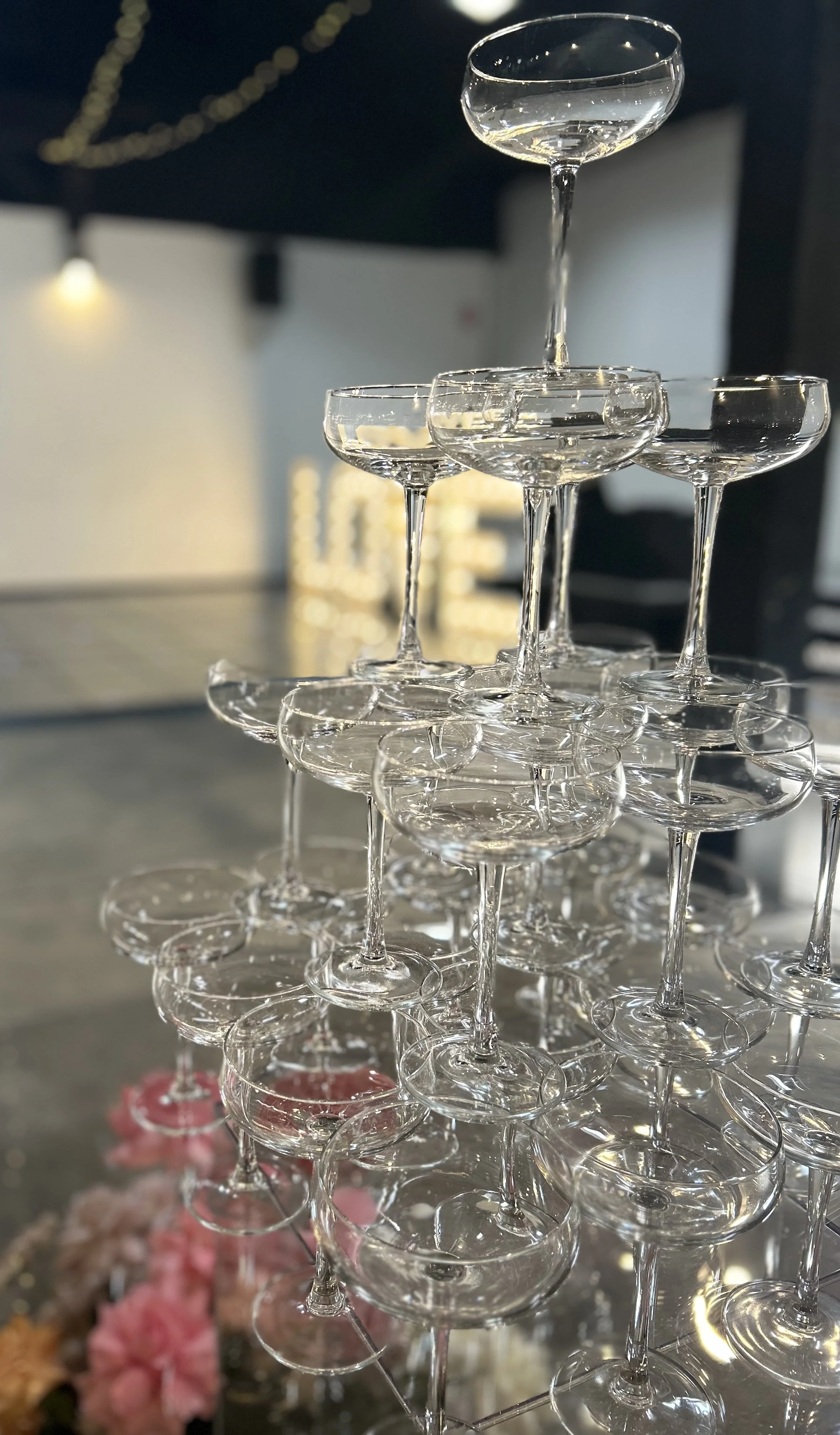 A pyramid of empty cocktail glasses stacked in a tower with a blurred background of lights and flowers.