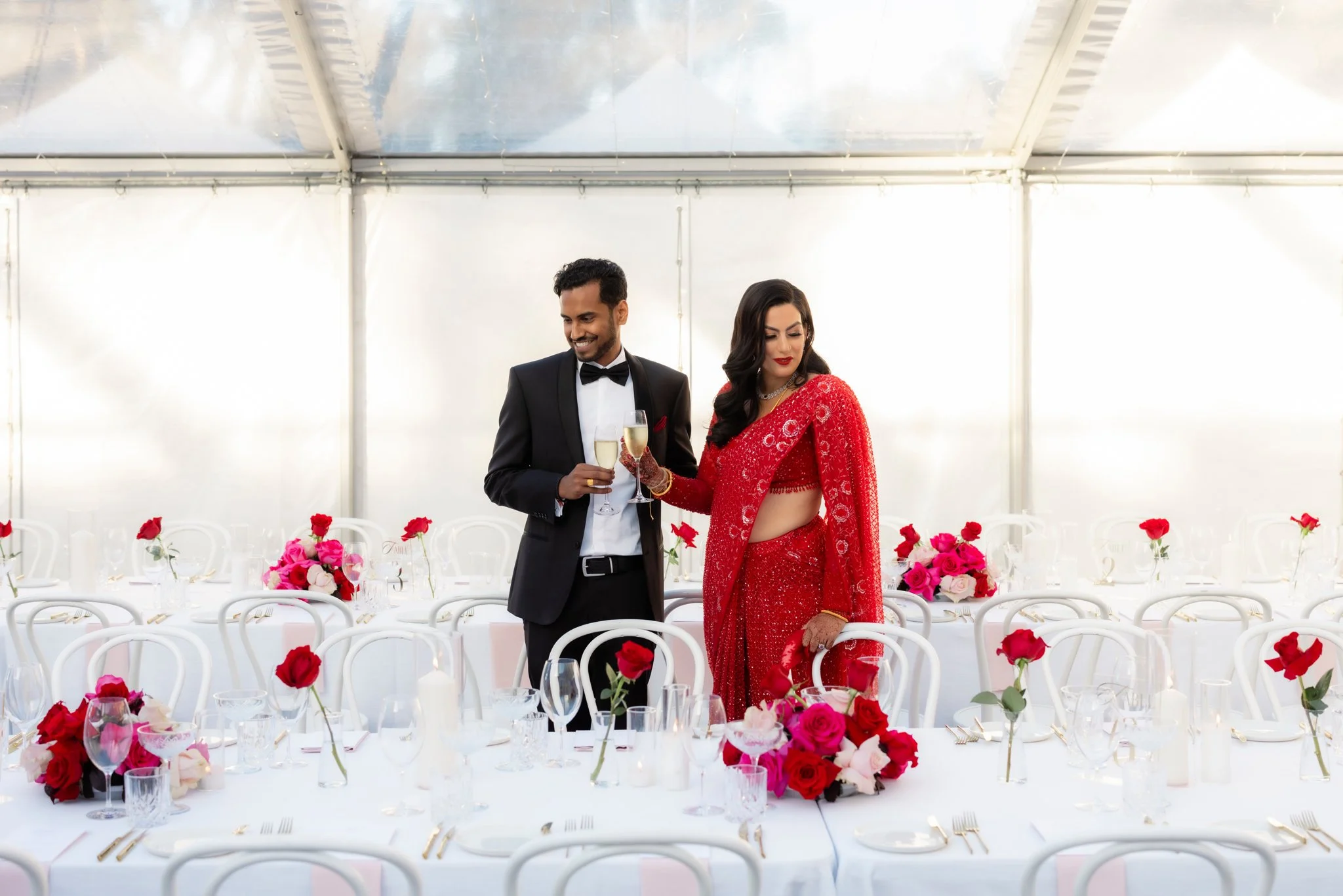 A man in a tuxedo and a woman in a red traditional outfit holding glasses of champagne at a decorated wedding reception table with pink and red flowers.