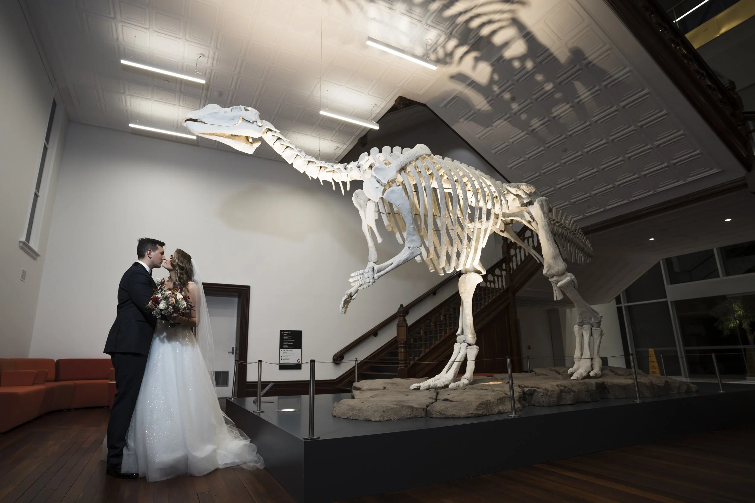 Bride and groom standing close, sharing a moment in front of a large dinosaur skeleton display in a museum.