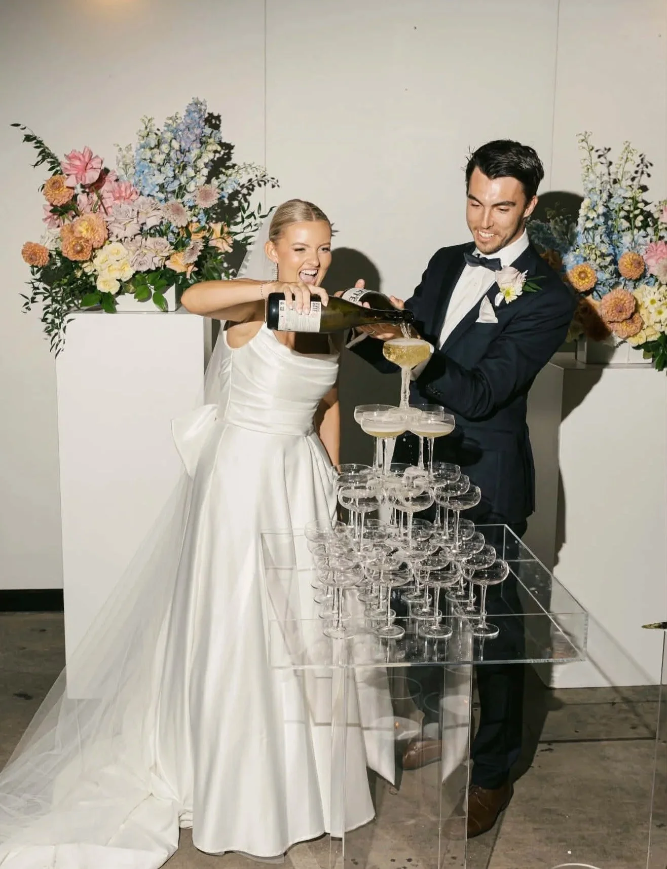 Bride and groom in wedding attire pouring champagne into a champagne tower, with large floral arrangements in the background.