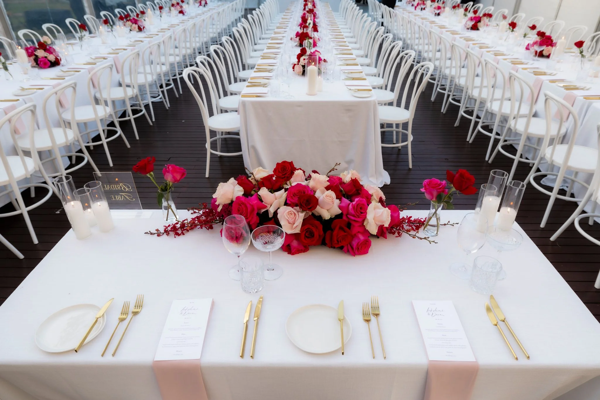 A long wedding reception table decorated with a floral centerpiece of red, pink, and white roses, with white candles in glass holders, surrounded by white chairs and a dark wooden floor.
