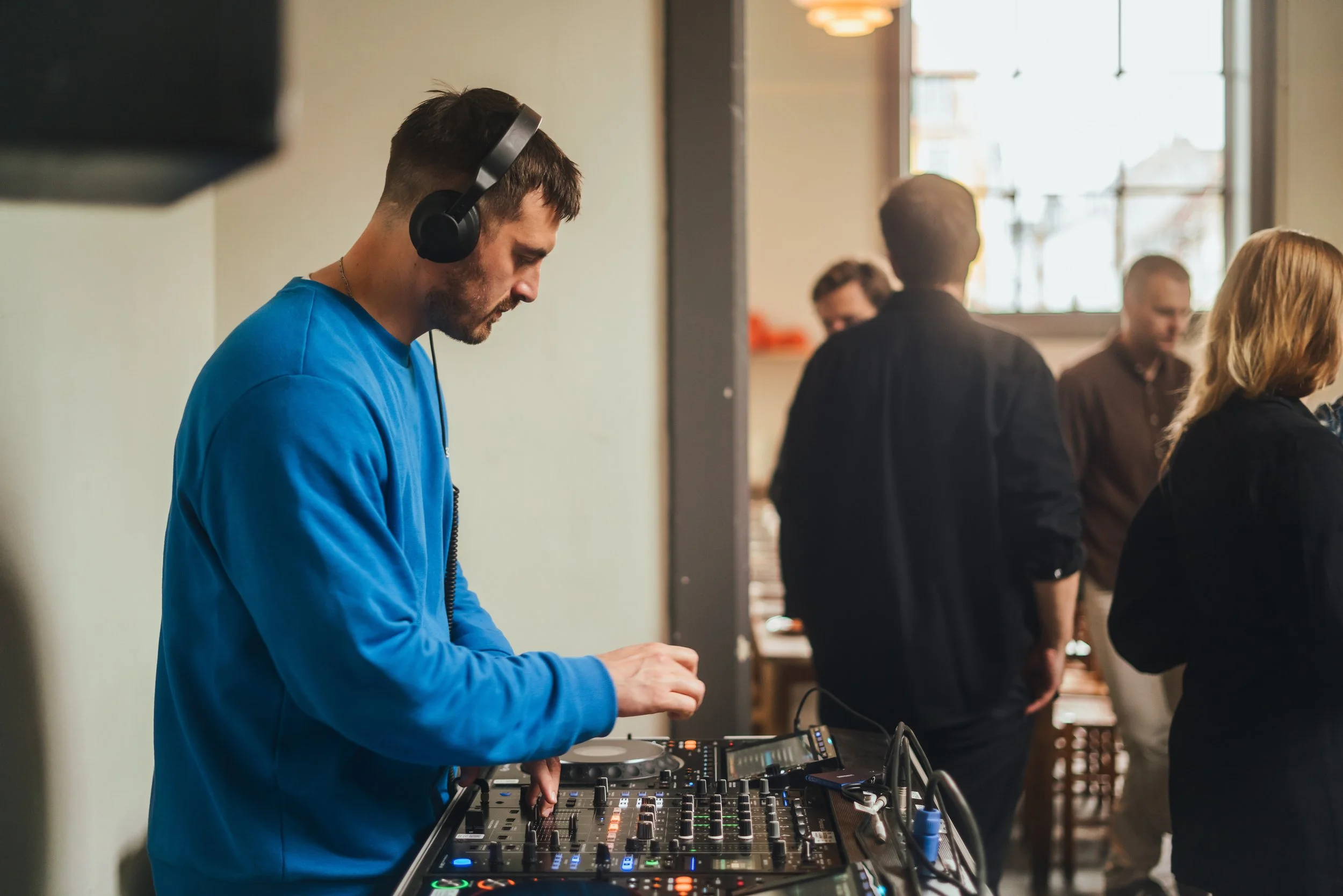 En mand i blå sweatshirt, med hovedtelefoner, DJ-udstyr og mixer, der spiller musik i et indendørs miljø med flere mennesker i baggrunden.