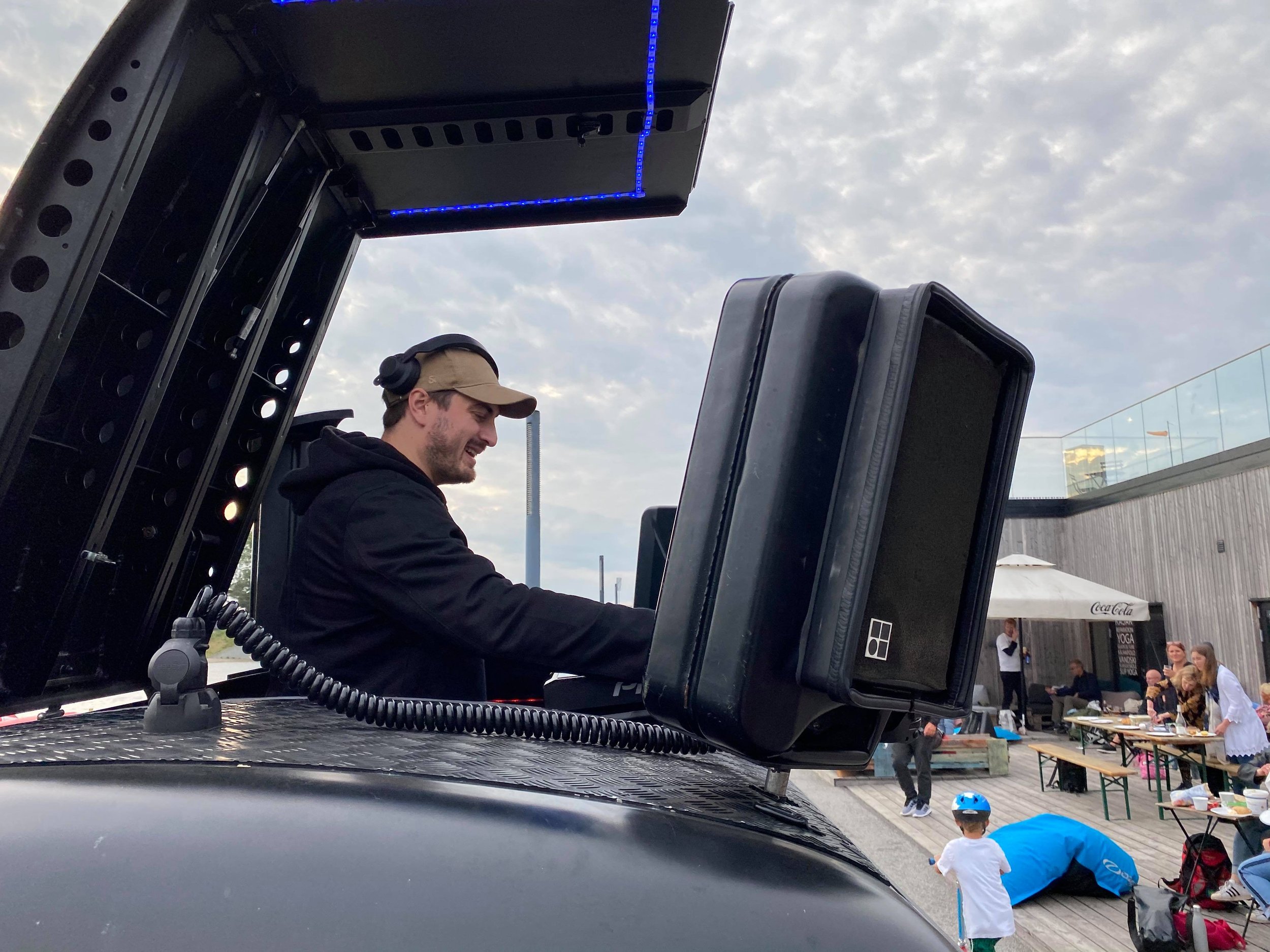 En mand med hovedtelefoner står bag en DJ-udstyr, med en stor skærm foran ham. I baggrunden er der et udeterrasse med mennesker, der står og sidder, nogle af dem med mad og drikke. En dreng med cykelhjelm går forbi.