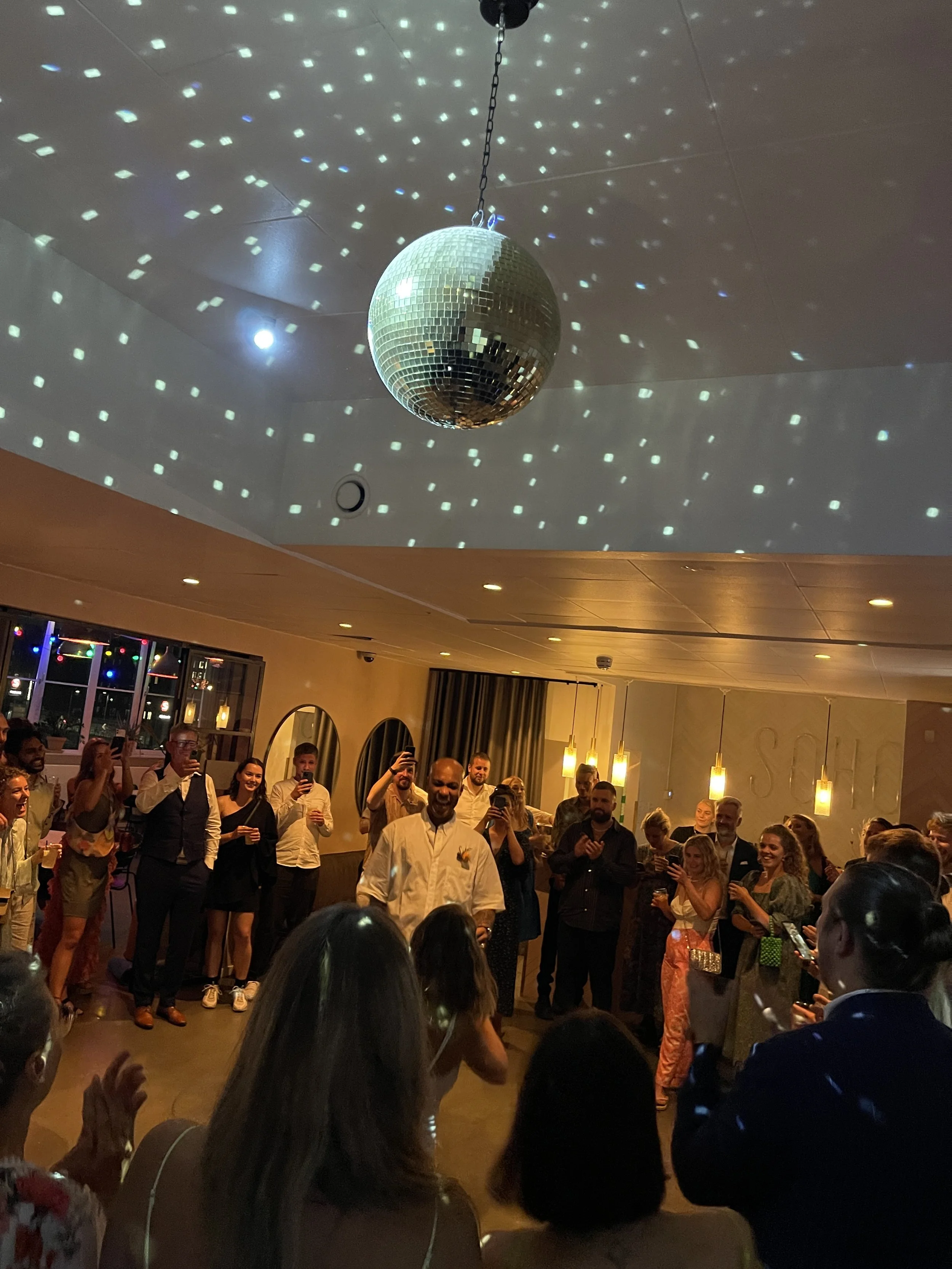 En gruppe mennesker står i en festlokale, hvor der er en diskokugle og lysreflekser i loftet. De ser ud til at nyde en festlig begivenhed, og der er mange smilende ansigter.