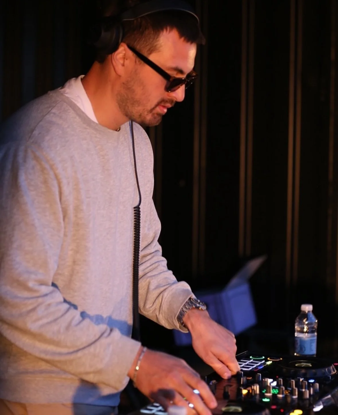 En mand, der DJ'er, står bag et DJ-udstyr med lysende knapper, i et mørkt rum, med en vandflaske på bordet.