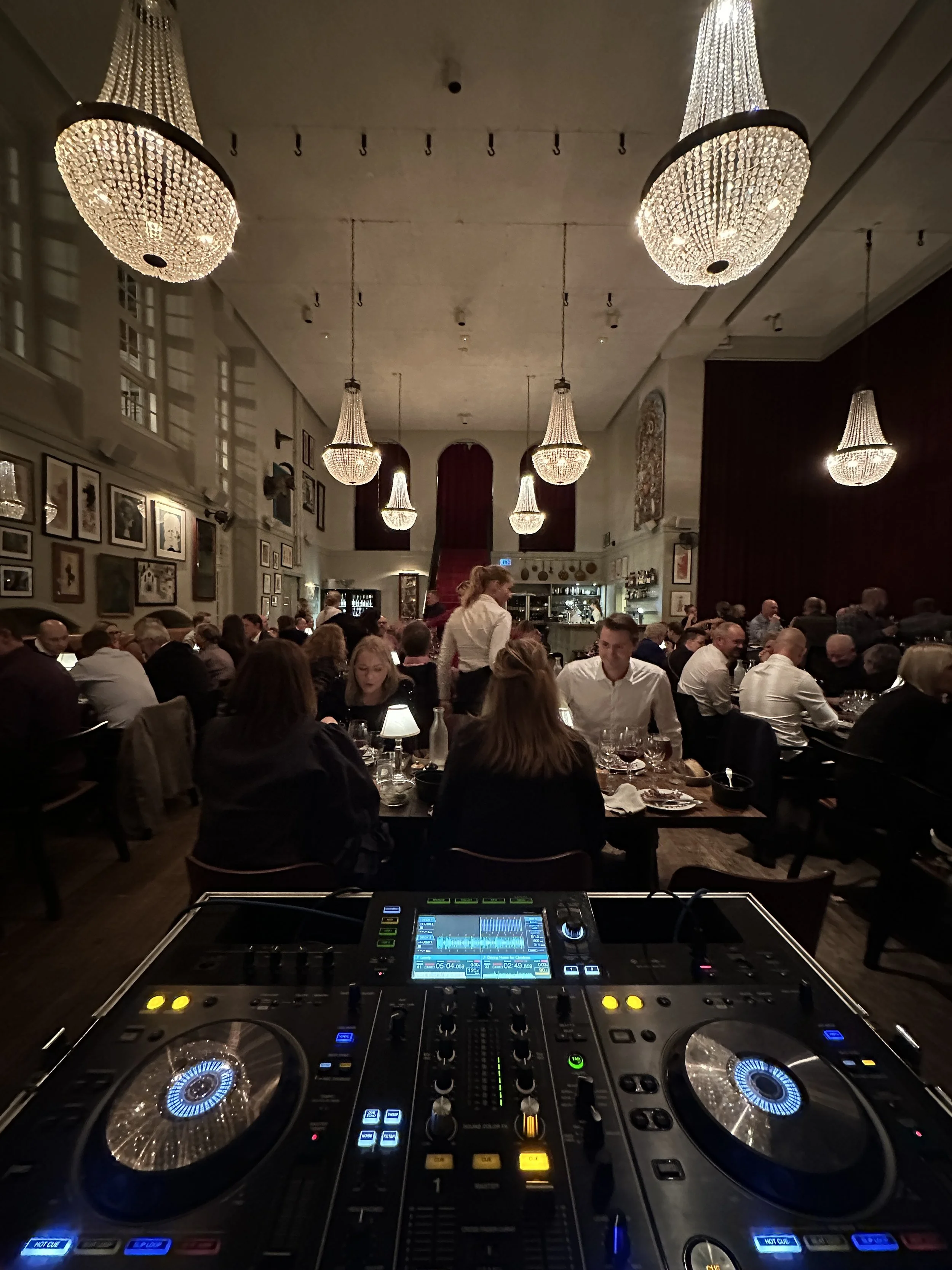 Billedet viser en elegant restaurant med mange gæster, dekorative lysekroner og vægkunst. I front er en DJ-setup med mixer og drejeskiver.