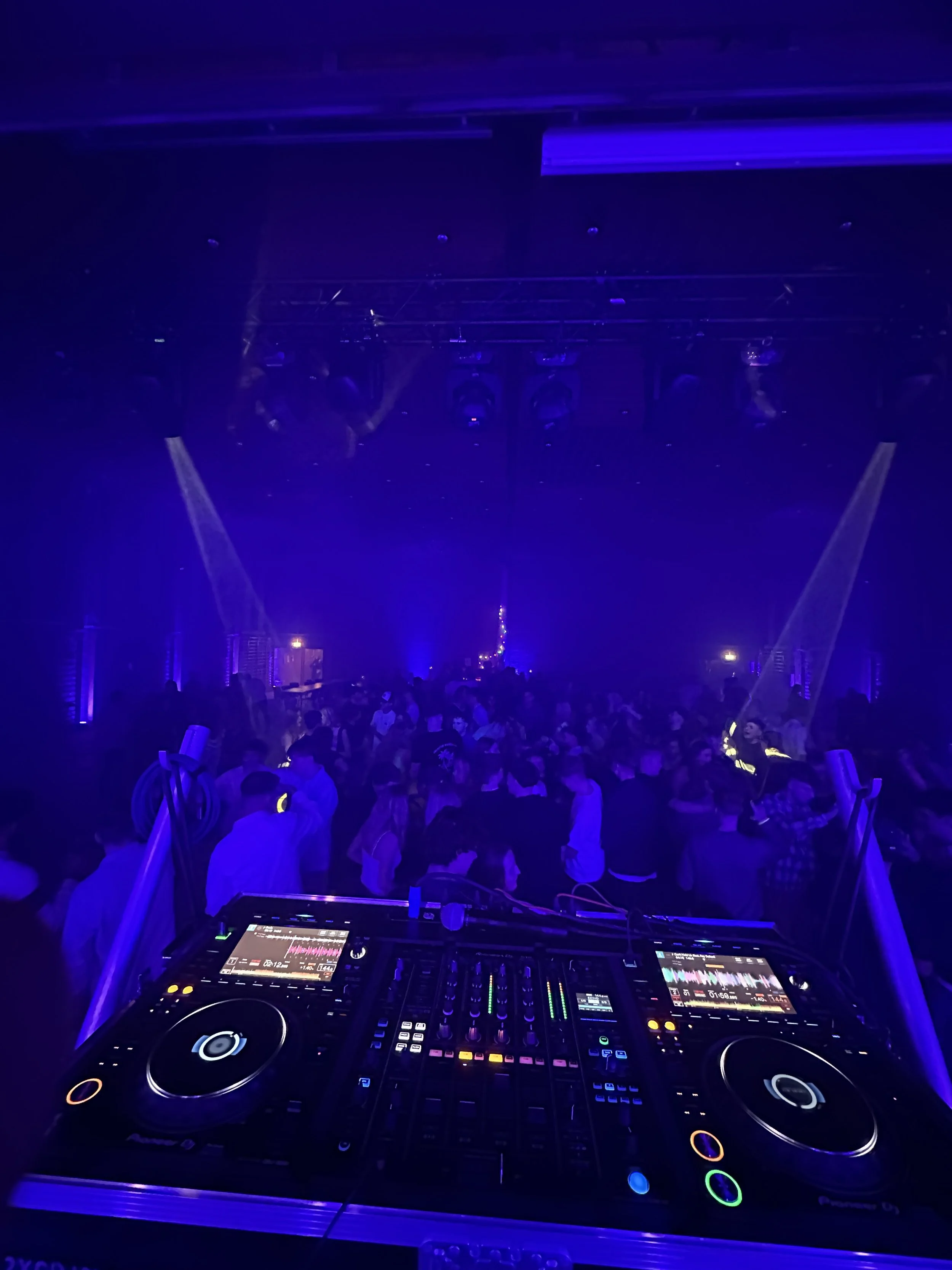 En DJ-spiller med udstyr i en nattelokale med blå belysning, og en stor dansefest med mange mennesker.