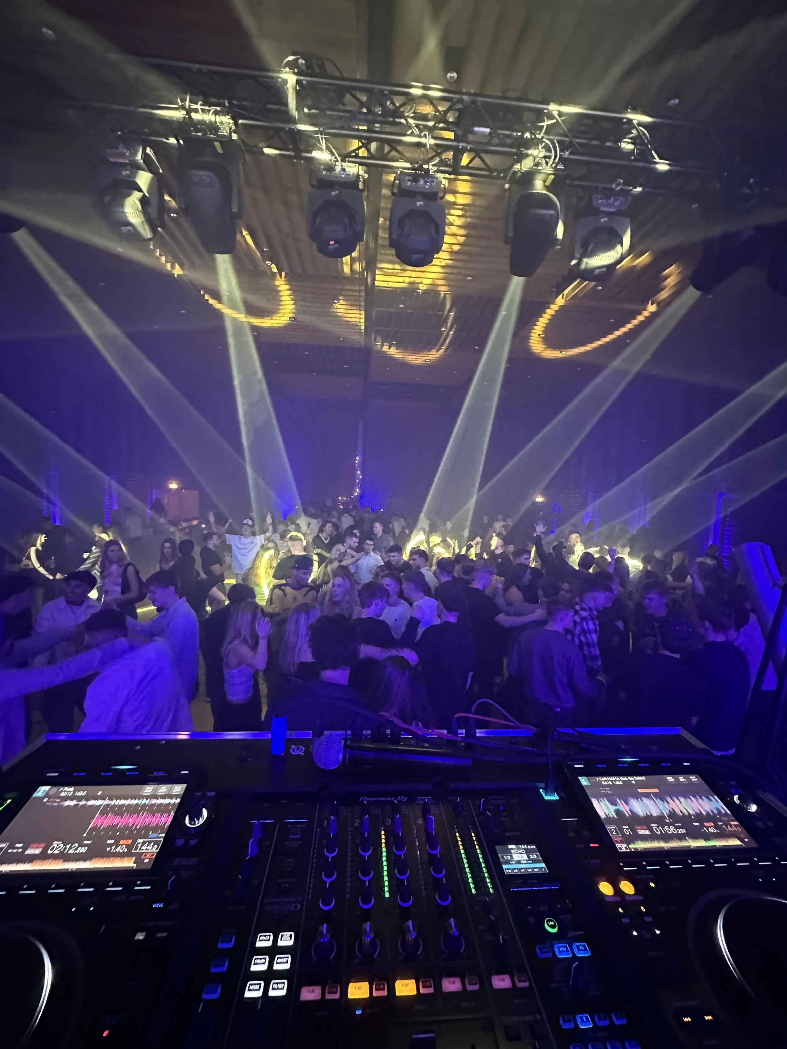 Et dansegulv fyldt med mennesker under en aftenfest med farvet lys og diskolys, set fra en DJ's vinkel med DJ-udstyr i forgrunden.