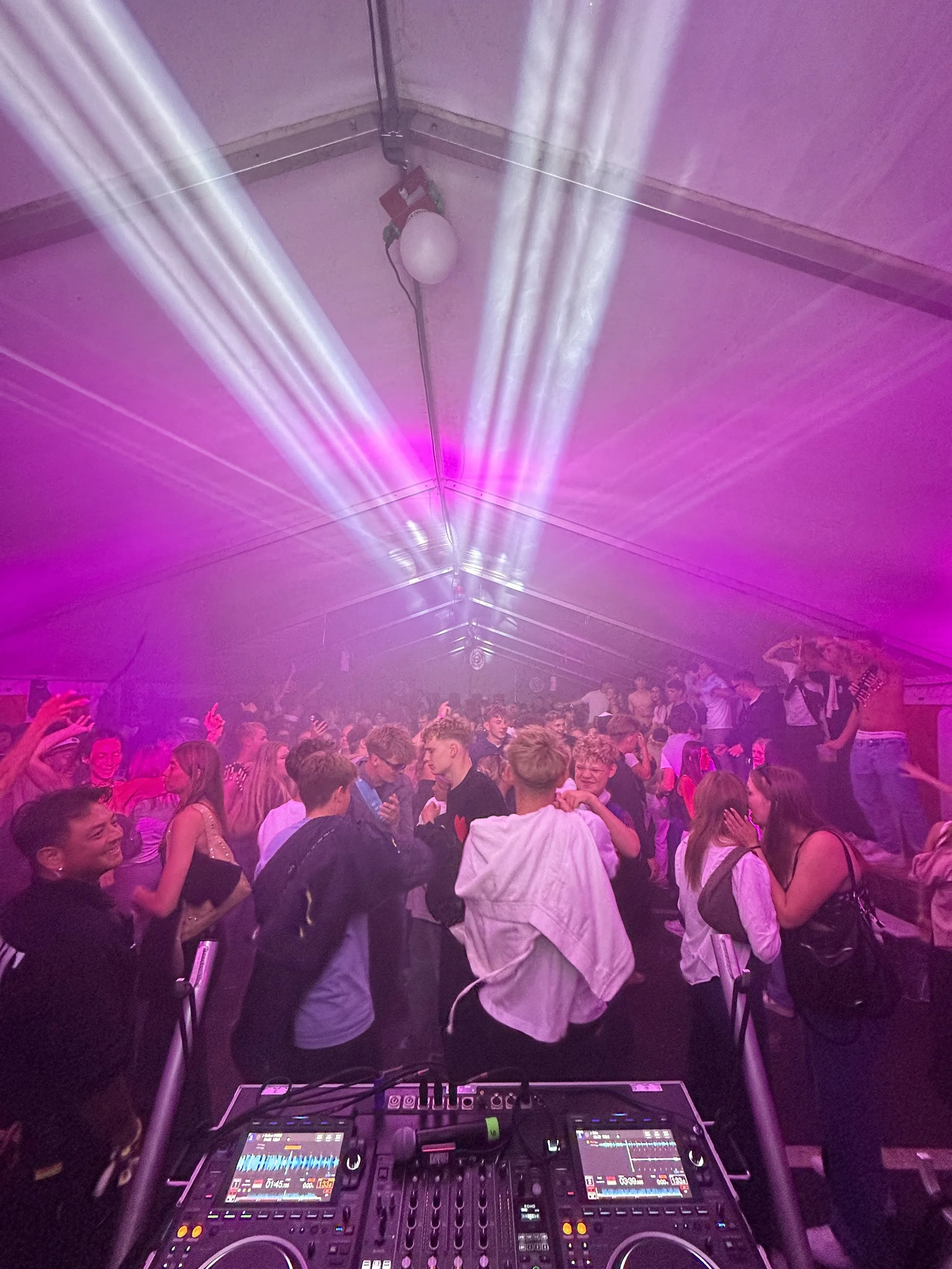 Dansende unge i en festtelt med belysning og DJ-udstyr i forgrunden.