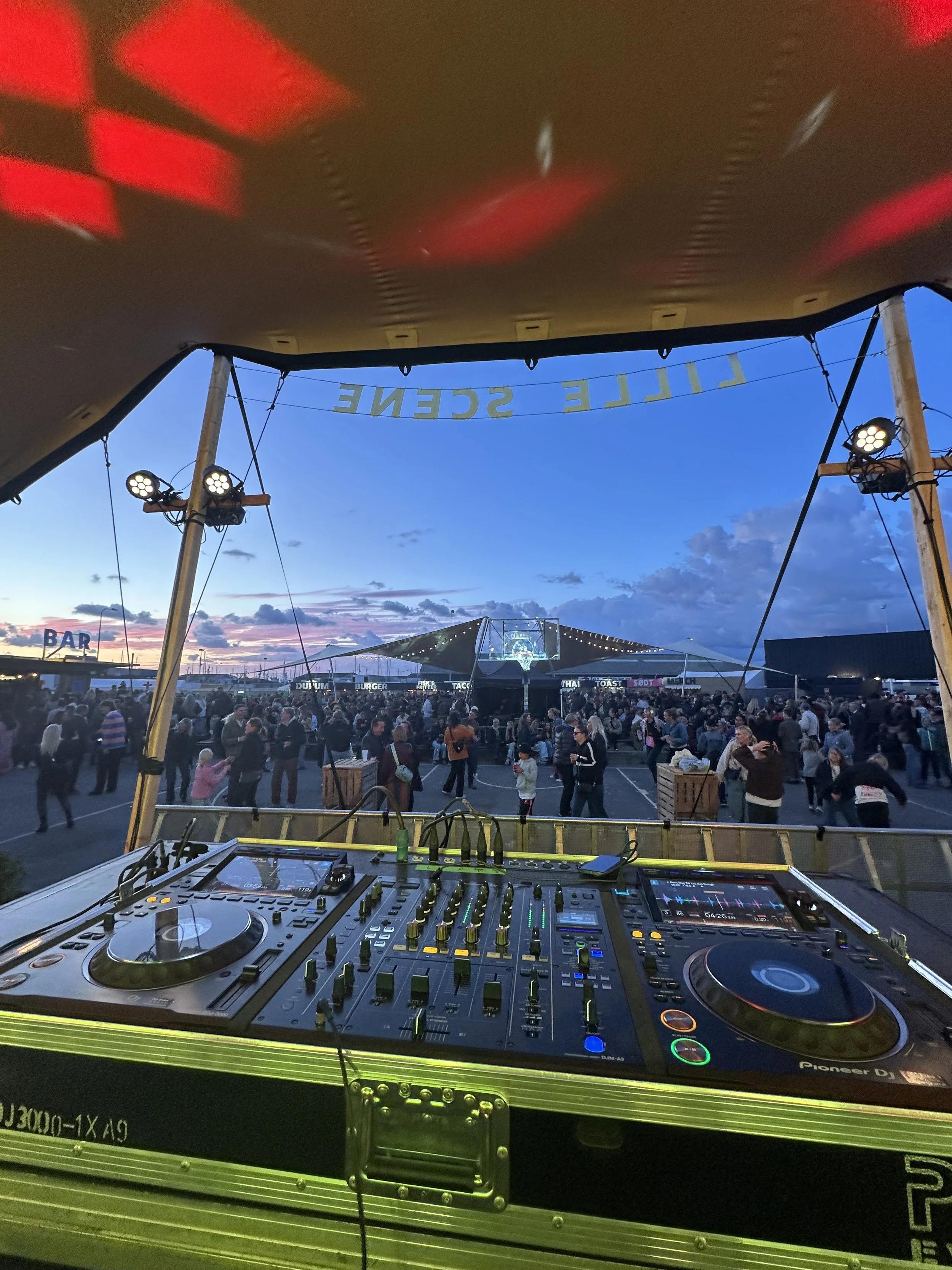 Udsigt over en festival med mange mennesker, et stort telt i baggrunden, en bar afskåret af lampelys, og en DJ-setup i forgrunden under en markise med teksten 'LILLE SCENE'.