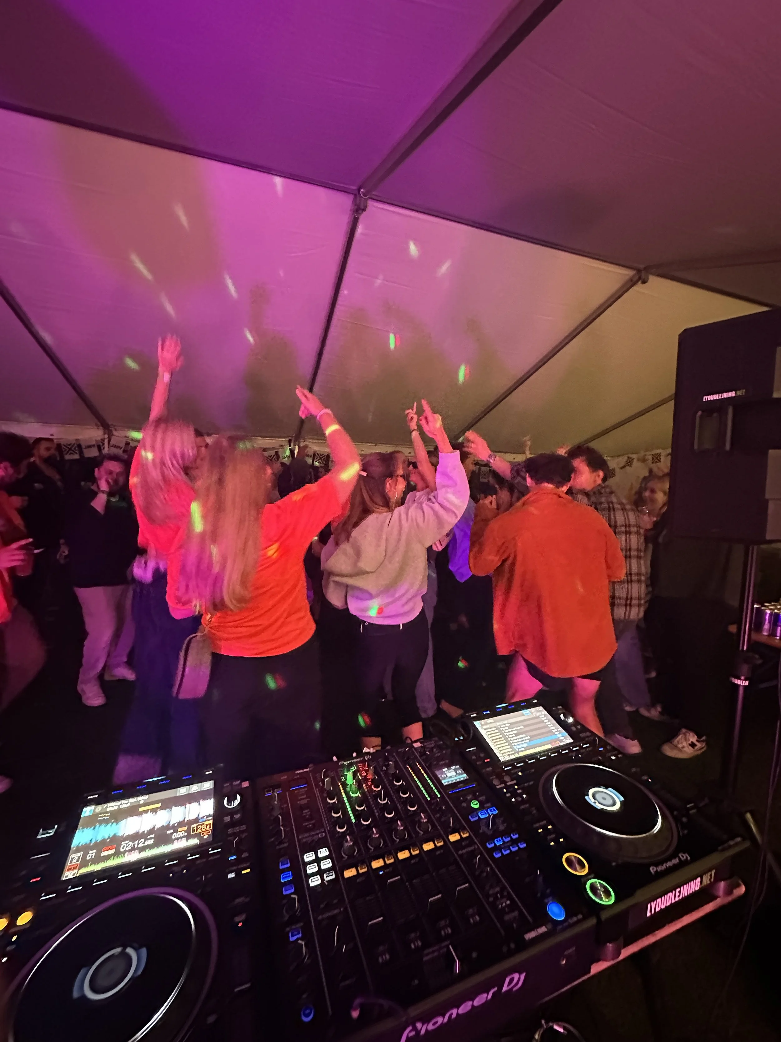 Folk danser og danser under en musikfestival i en stor telt med farvet belysning, foran en DJ-setup.