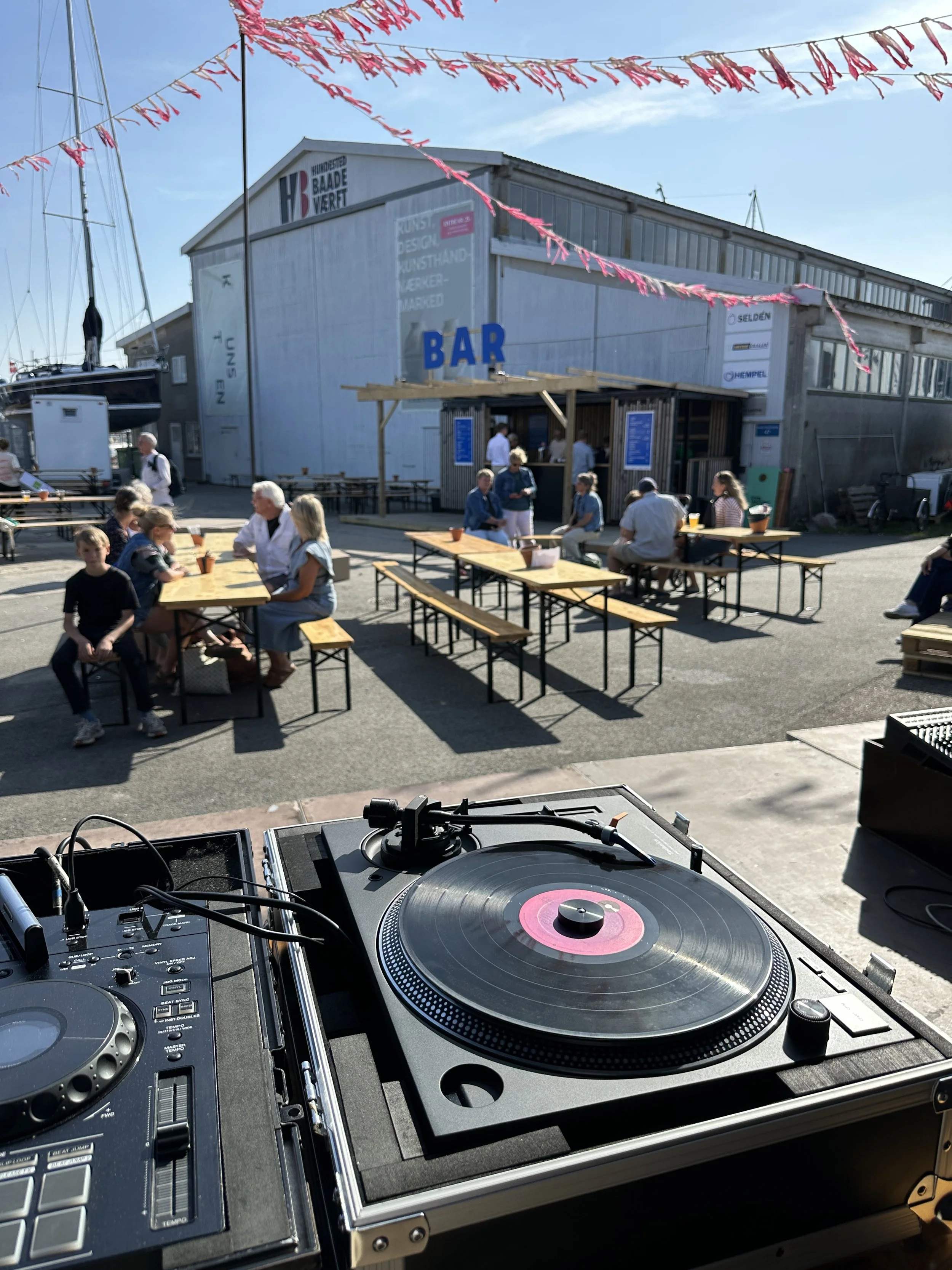 Udendørs scene ved marina med mennesker, sidder ved borde, der er en bar bygget af træ foran en stor lagerbygning, pink flagranker hænger over, DJ-udstyr i forgrunden, på en solrig dag.