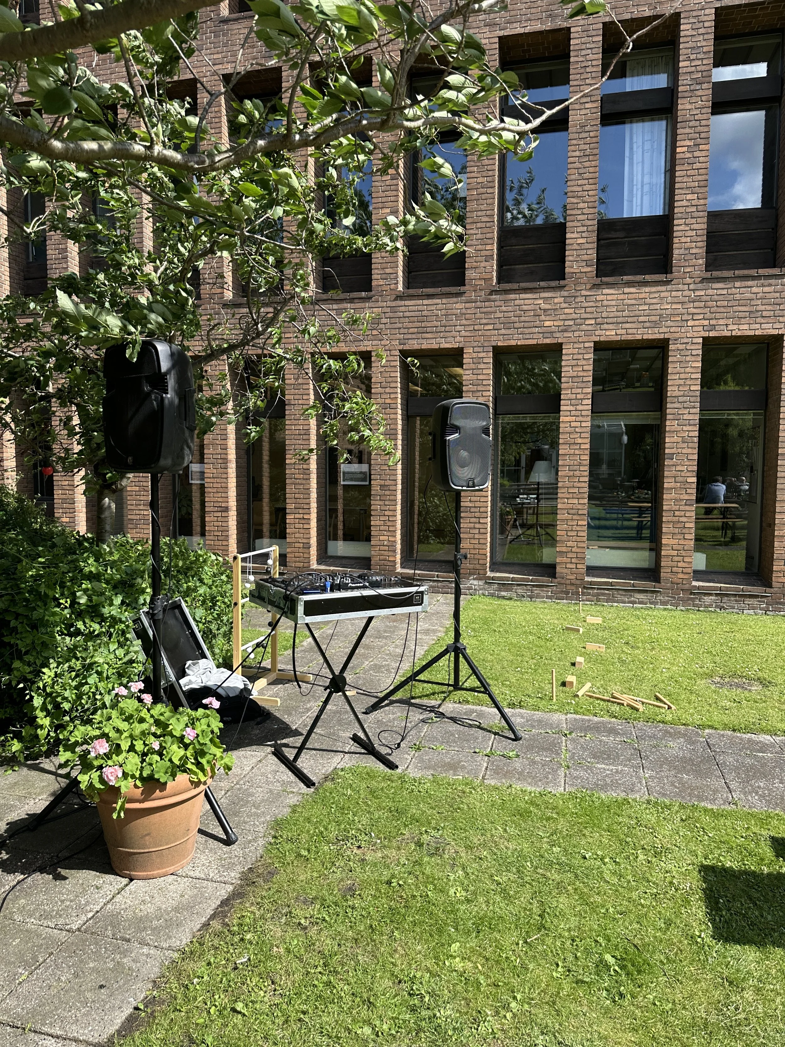 DJ-udstyr sat op udenfor foran en murstensbygning med store vinduer, omgivet af grønne planter og græs, under en trækane.