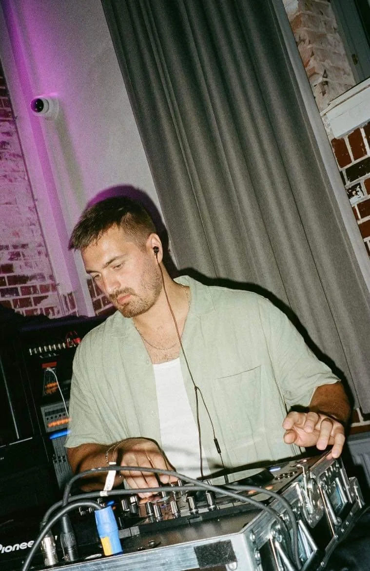 En mand, der spiller på en DJ-controllermaskine med høretelefoner, i et indendørs område med gardiner og murstensvæg.