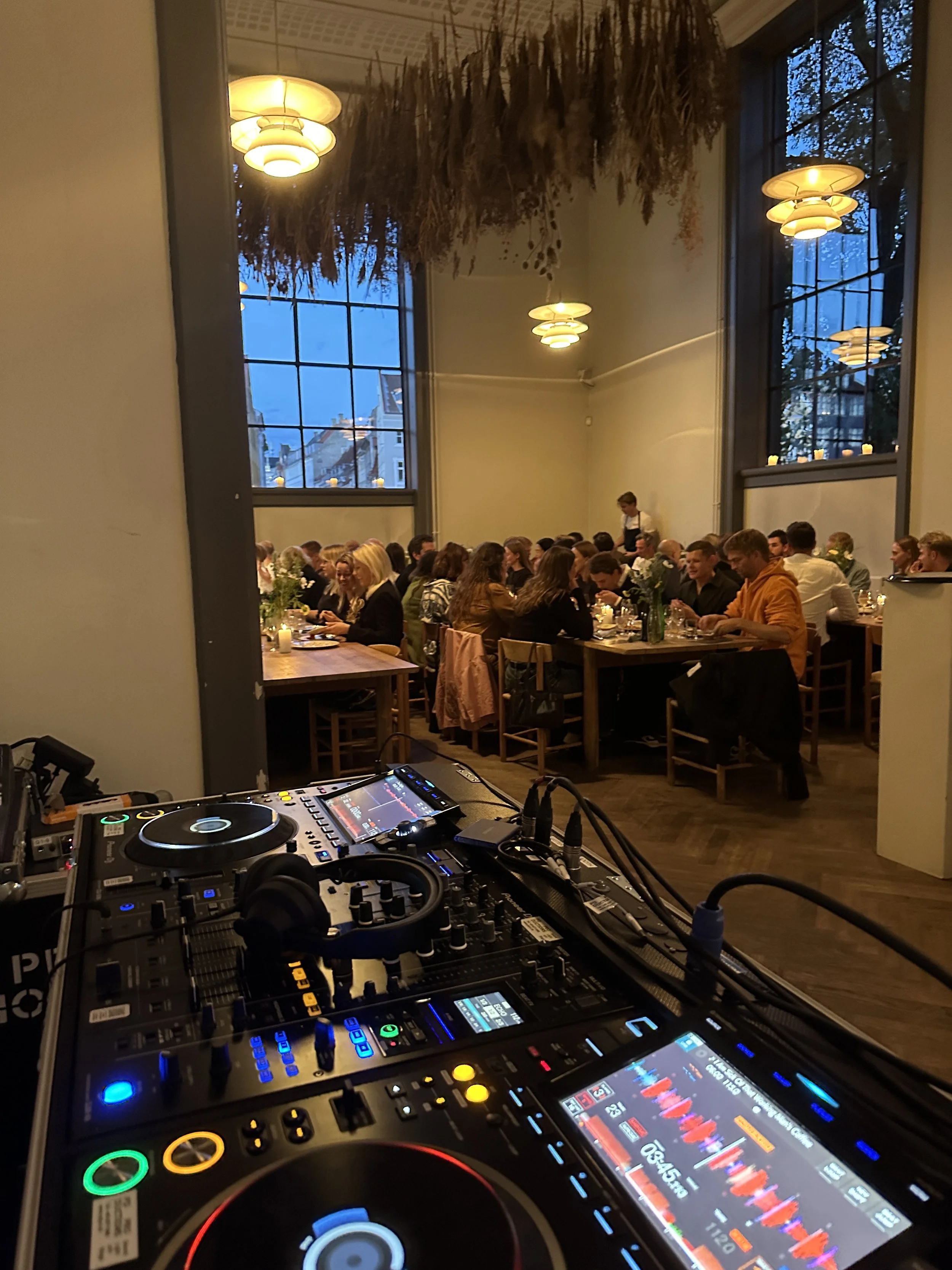 Indendørs restaurant med mange mennesker siddende ved langbord, der spiser og taler, taget fra DJ-udstyret set!i forgrunden