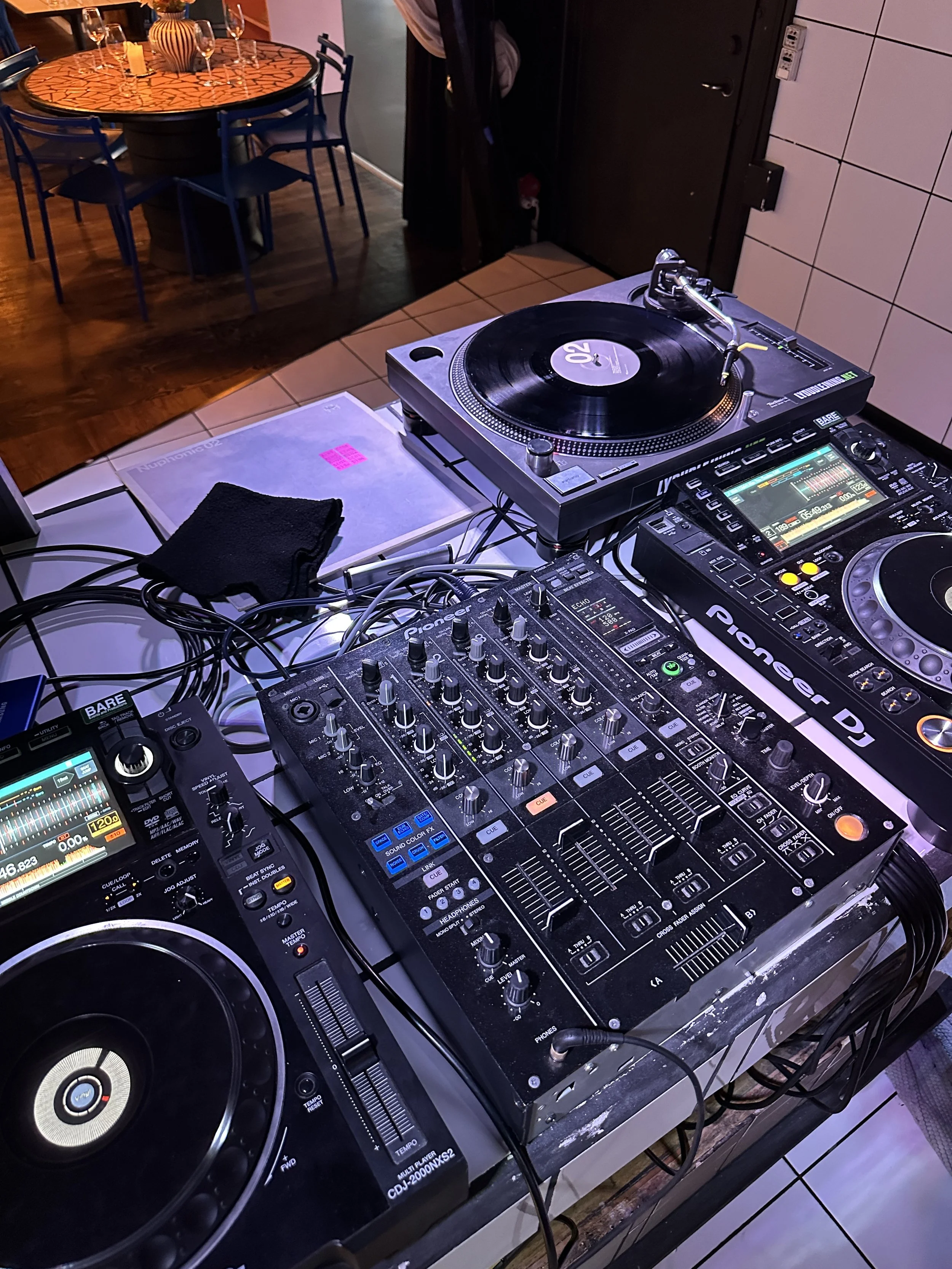 DJ-udstyr med to cd-afspillere, en mixer, og en DJ-controller, sat op på et bord i en restaurant eller klub.