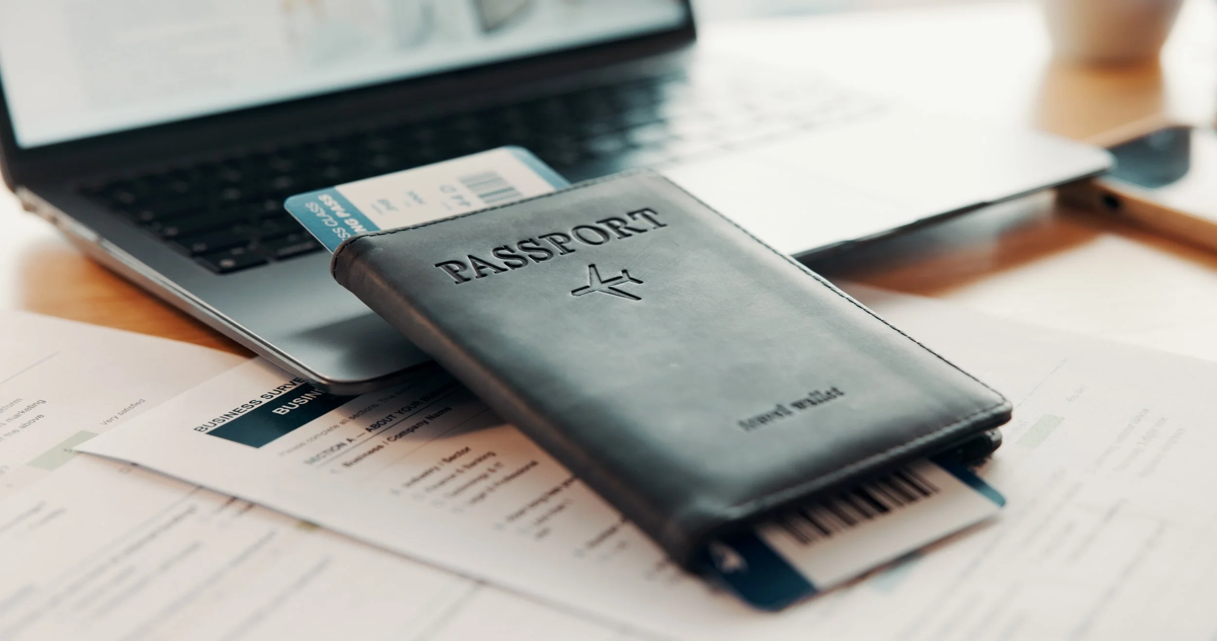 Laptop with a passport holder, boarding pass and business travel documents on a desk.