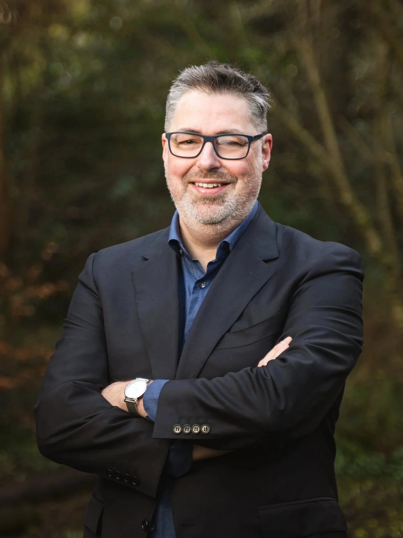 A man with gray hair and beard wearing glasses, a black blazer, and a blue shirt, standing outdoors with arms crossed in front of a blurred natural background. Alan Athey