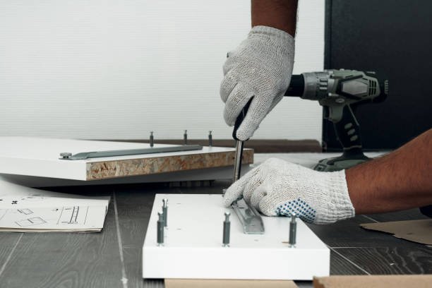 Person montiert Holzplatten mit Schrauben auf einer Arbeitsplatte, trägt Arbeitshandschuhe und benutzt ein** Akkuschrauber. Im Hintergrund liegt eine weiteres Holzstück.